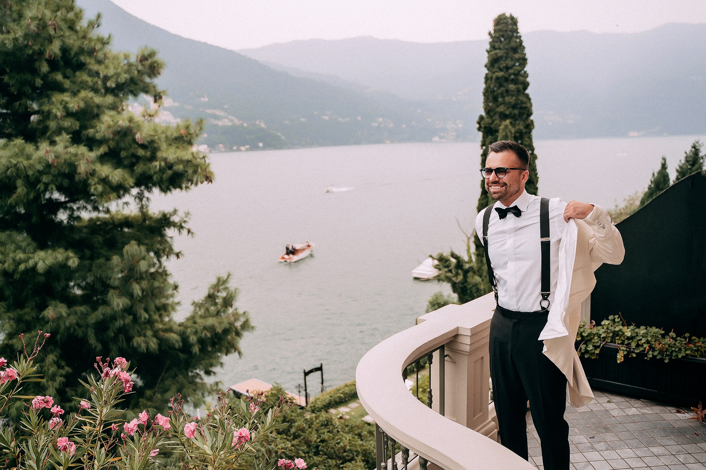 The groom in formal attire—stands on a balcony holding a beige jacket. Behind them, lush greenery and pink flowers frame a tranquil lake with boats and hazy mountain views.