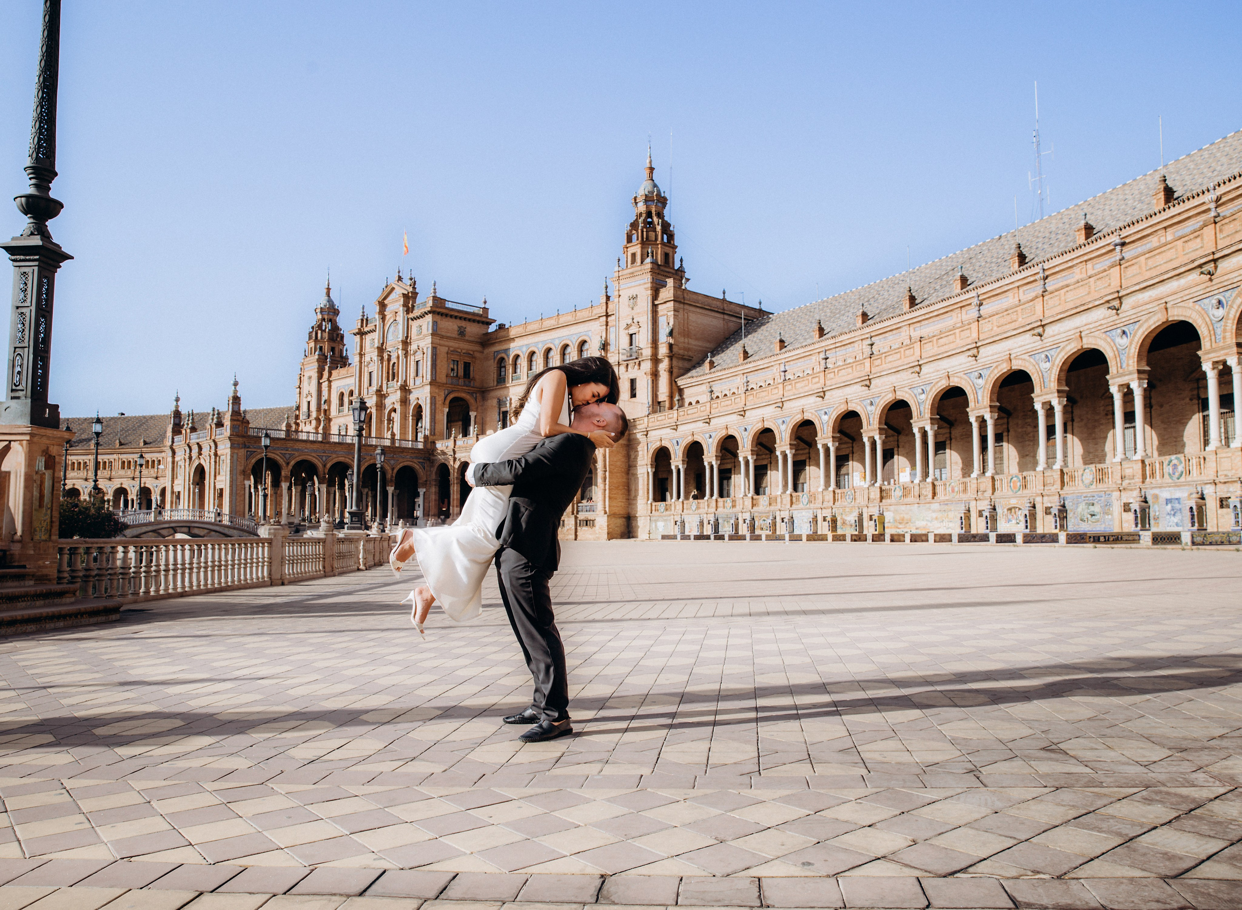Joyful wedding photo of a groom lifting his bride in front of the iconic Plaza de España in Sevilla, Spain — perfect inspiration for romantic and cinematic wedding photoshoots in Seville.
