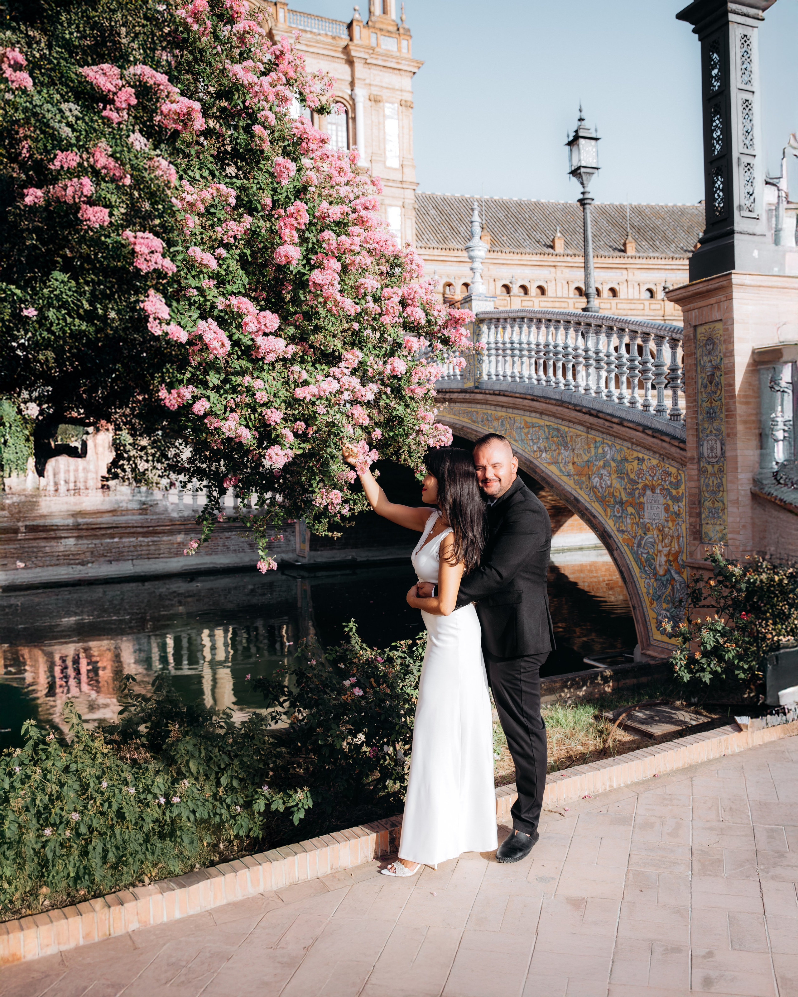 Romantic wedding photo in Sevilla, Spain, showing a couple embracing under blooming pink flowers near the iconic ceramic bridge at Plaza de España — perfect inspiration for couples searching for a floral wedding photoshoot in Seville or destination wedding photography in Spain.