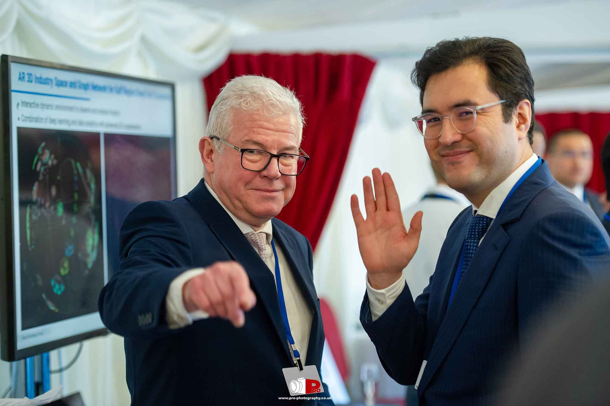 Two businessmen smiling and greeting attendees with a wave at a corporate networking event.