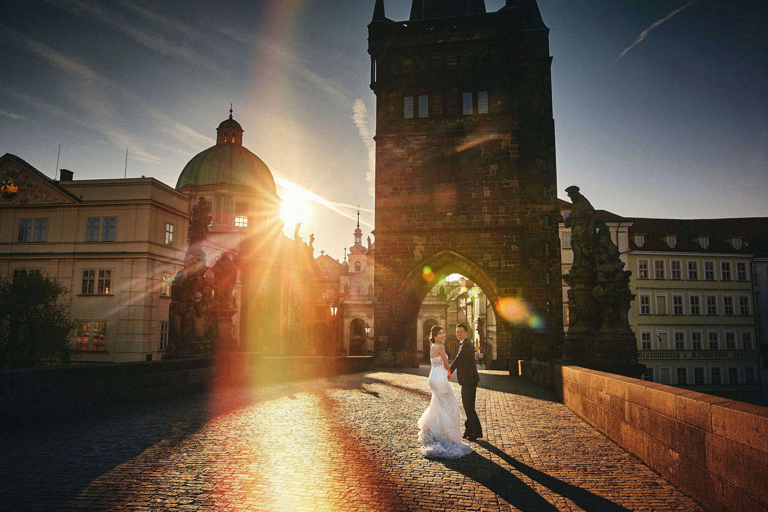 Smiling Hong Kong bride looking back while running with groom towards Charles Bridge tower under a vivid sun flare.
