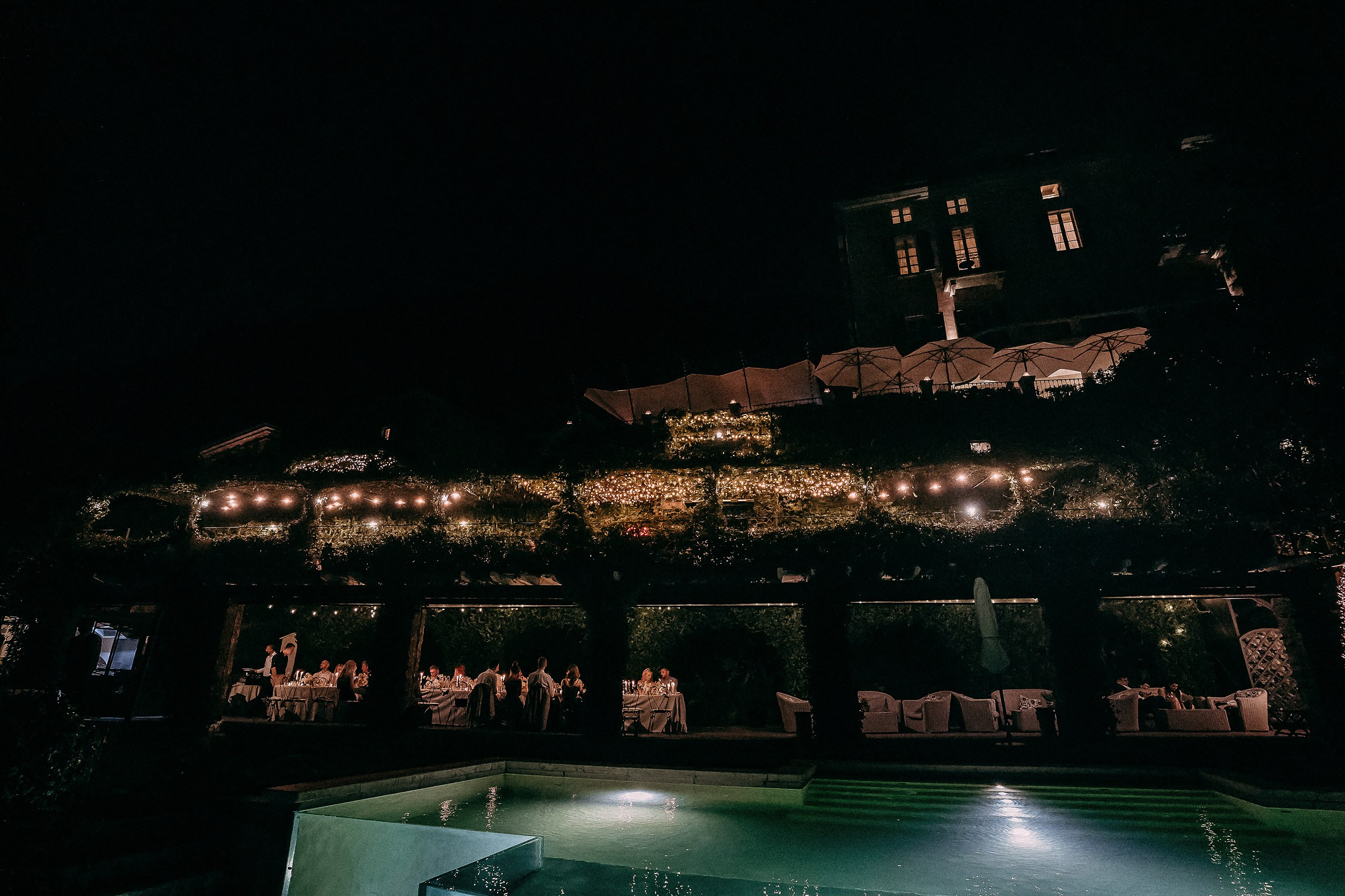 Romantic nighttime wedding reception on terrace with string lights, lush greenery, and pool reflections — guests dining under umbrellas in warm, ambient glow for luxury outdoor event photography.