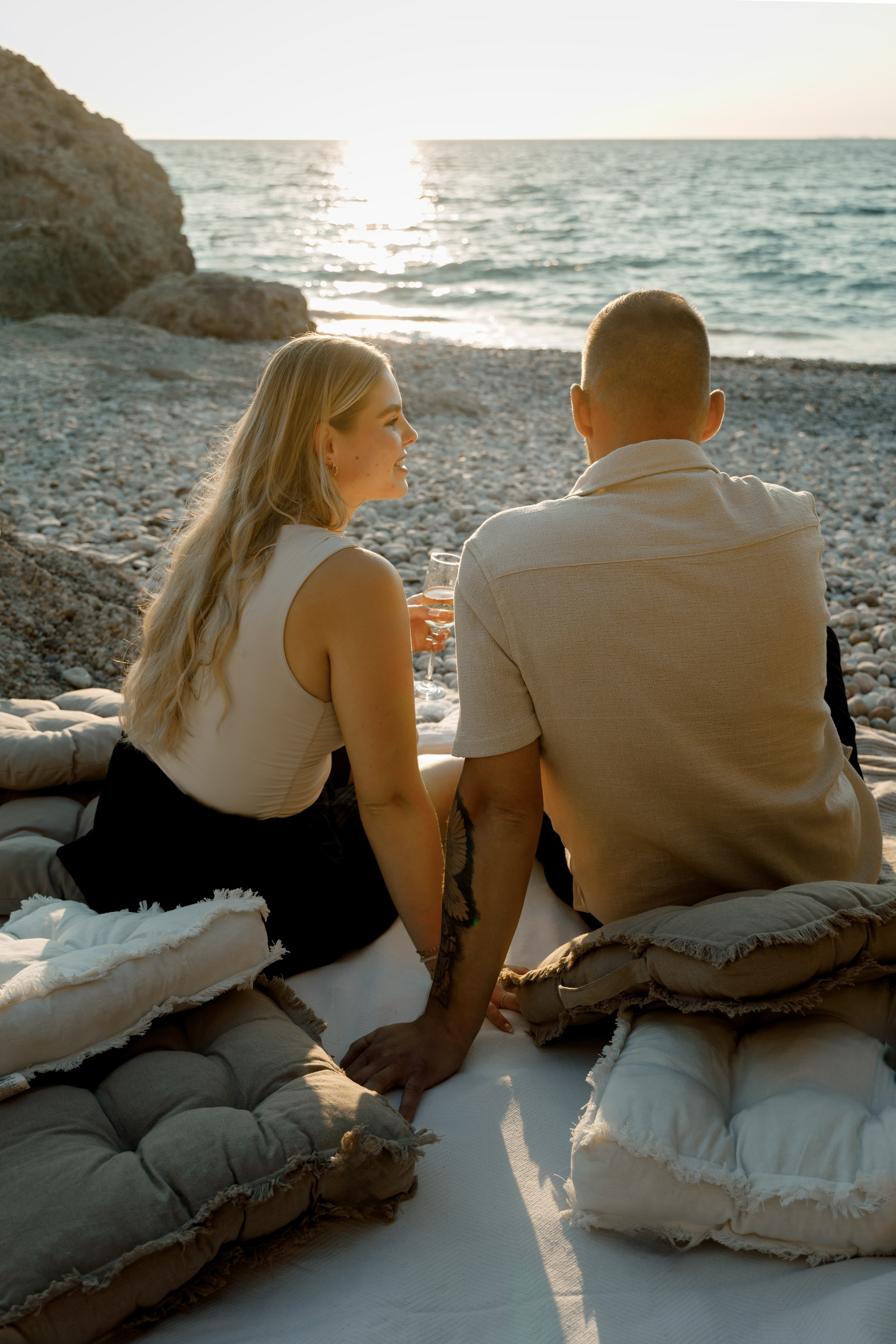 "Moment der Ruhe nach dem Ja-Wort: Ein Paar sitzt auf dem Kieselstrand von Kato Petres und genießt die Zweisamkeit. Die Wellen schlagen sanft an das steinige Ufer, und das warme Licht des Sonnenuntergangs taucht die Szenerie in goldene Farben."