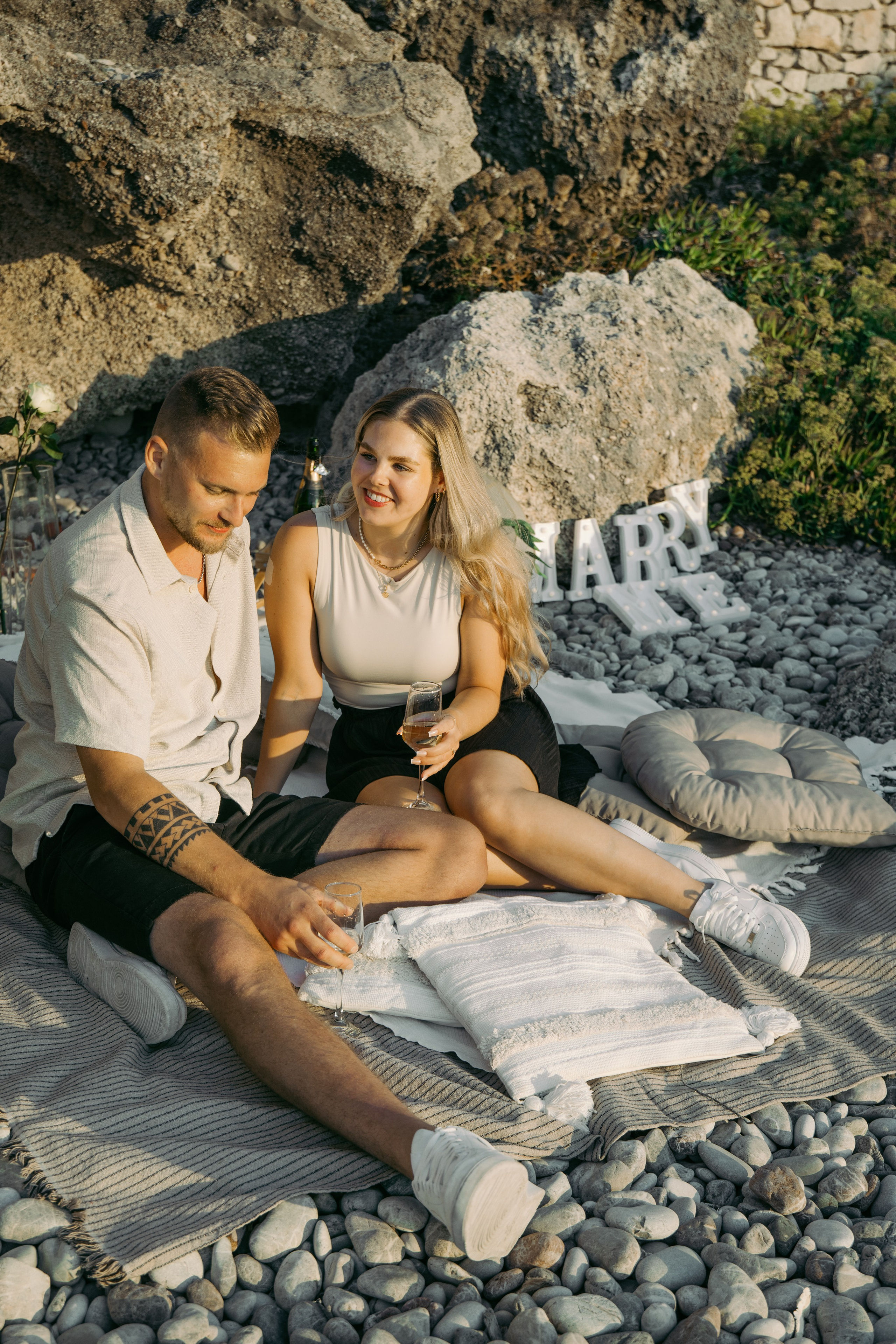 "Moment der Ruhe nach dem Ja-Wort: Ein Paar sitzt auf dem Kieselstrand von Kato Petres und genießt die Zweisamkeit. Die Wellen schlagen sanft an das steinige Ufer, und das warme Licht des Sonnenuntergangs taucht die Szenerie in goldene Farben."