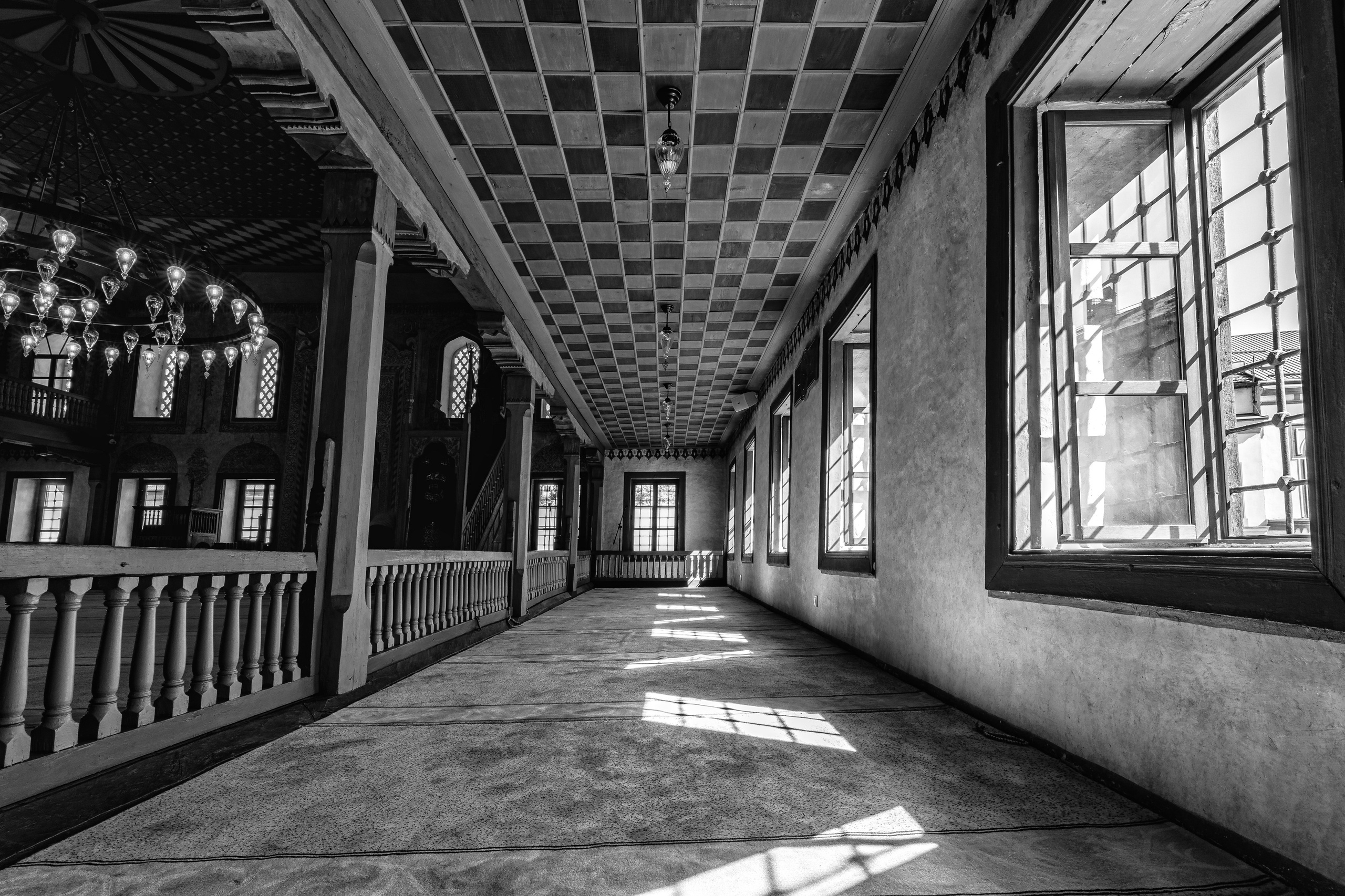 Interior architectural photography of a historic Ottoman mosque featuring geometric ceiling patterns, arched windows, wooden galleries and decorative chandeliers illuminated by natural light.