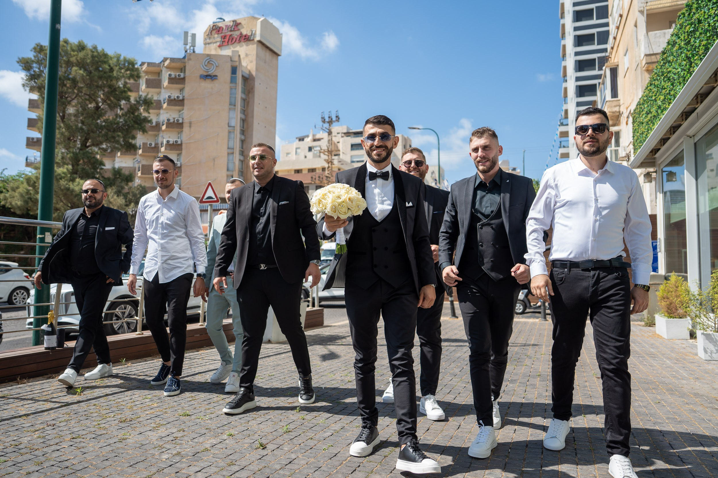 Groom Shimon holding a white rose bouquet and walking down the street with his groomsmen in Netanya, Israel
