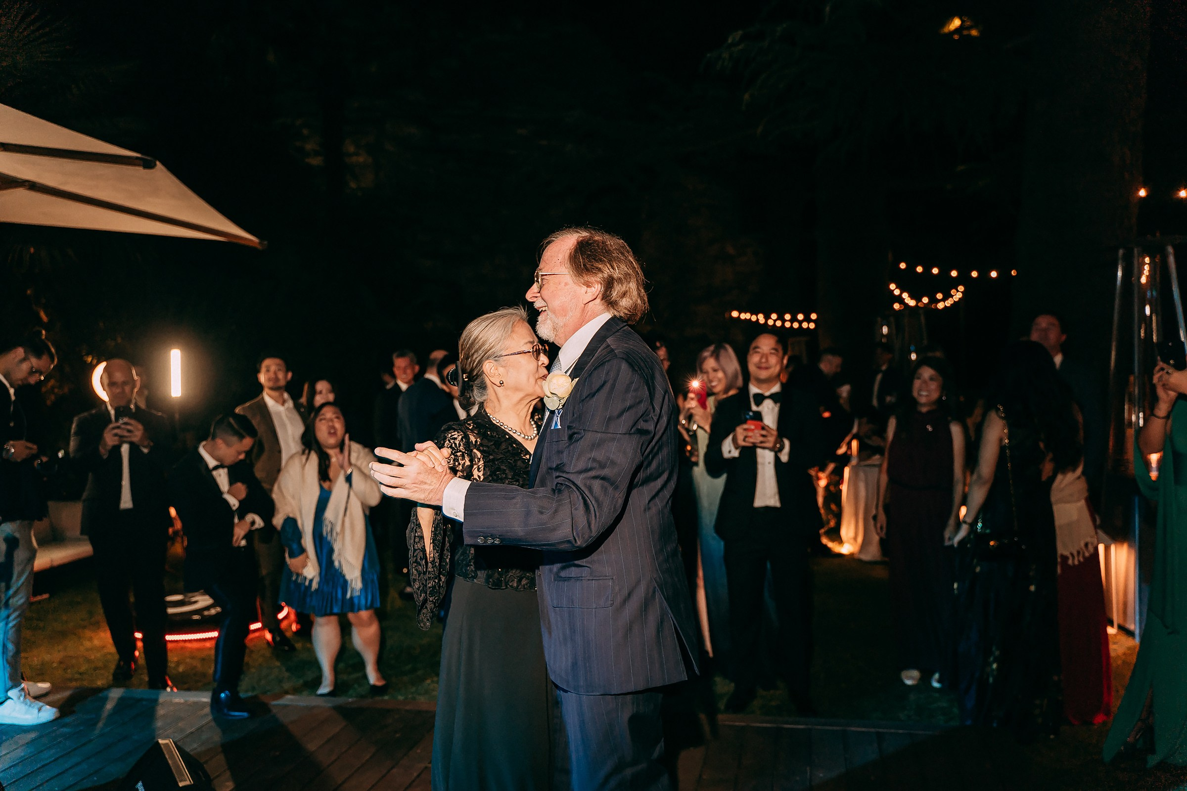 Elderly couple dances closely, surrounded by joyful guests in a warmly lit outdoor wedding celebration.