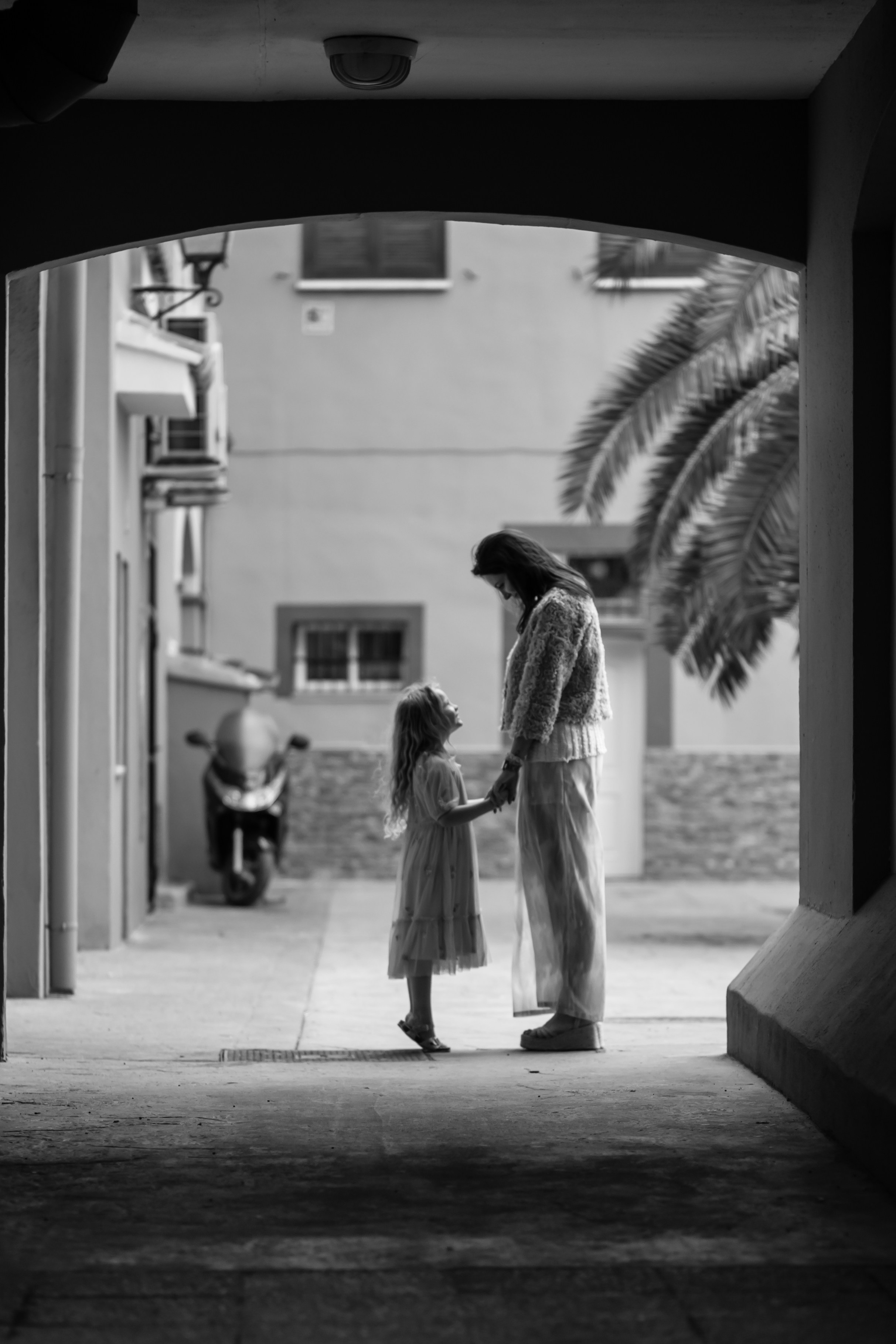 Retrato familiar atemporal en blanco y negro de una madre e hija compartiendo un momento tranquilo bajo un arco en un encantador callejón de Valencia, España — capturando un lazo entrañable y la belleza de lo cotidiano.