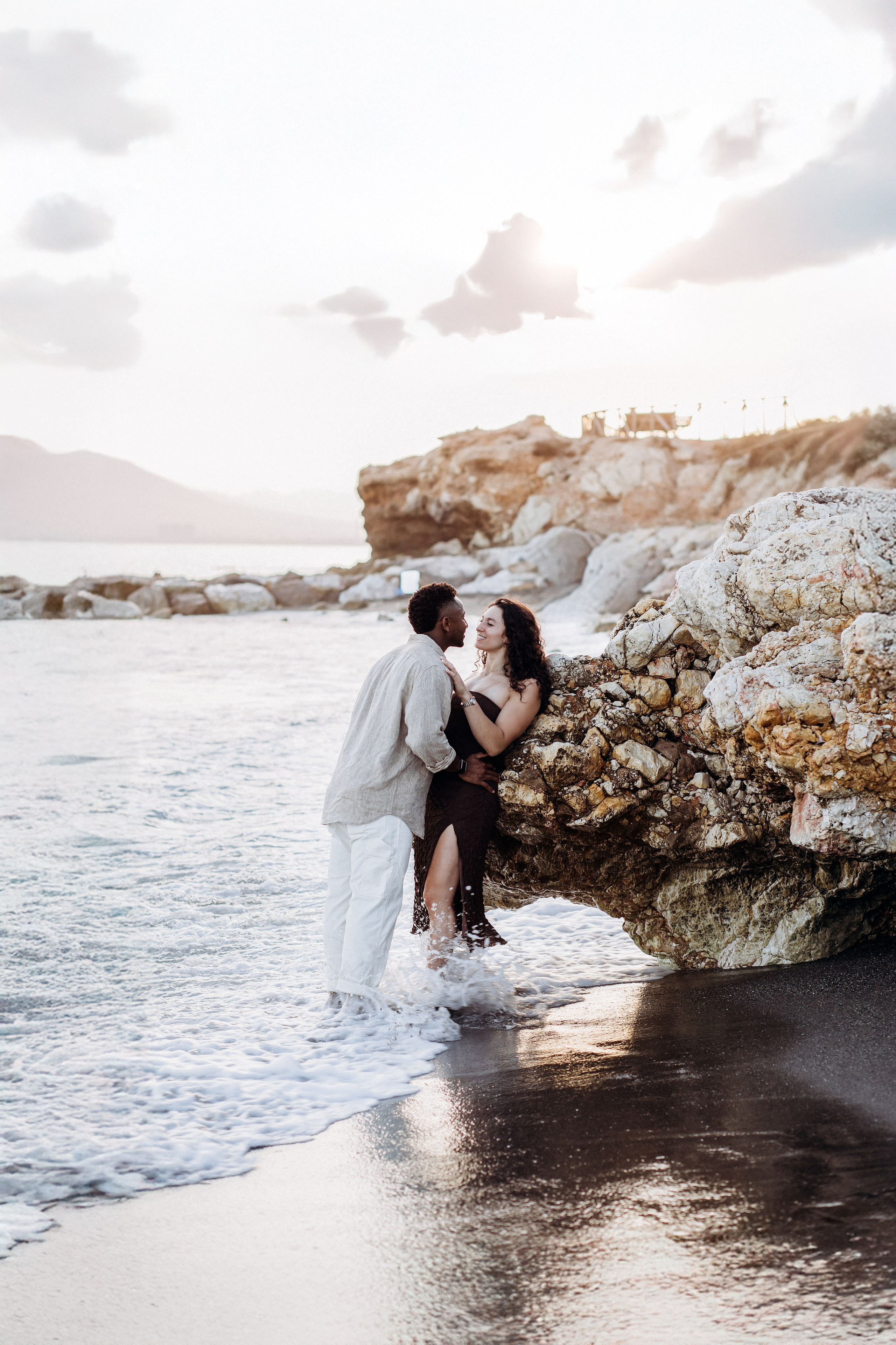 Pareja romántica sentada junto a las rocas al borde del agua durante una sesión de historia de amor a la hora dorada en Valencia, España — perfecto para parejas que buscan sesiones espontáneas y emocionales en la playa de Valencia y en toda España.