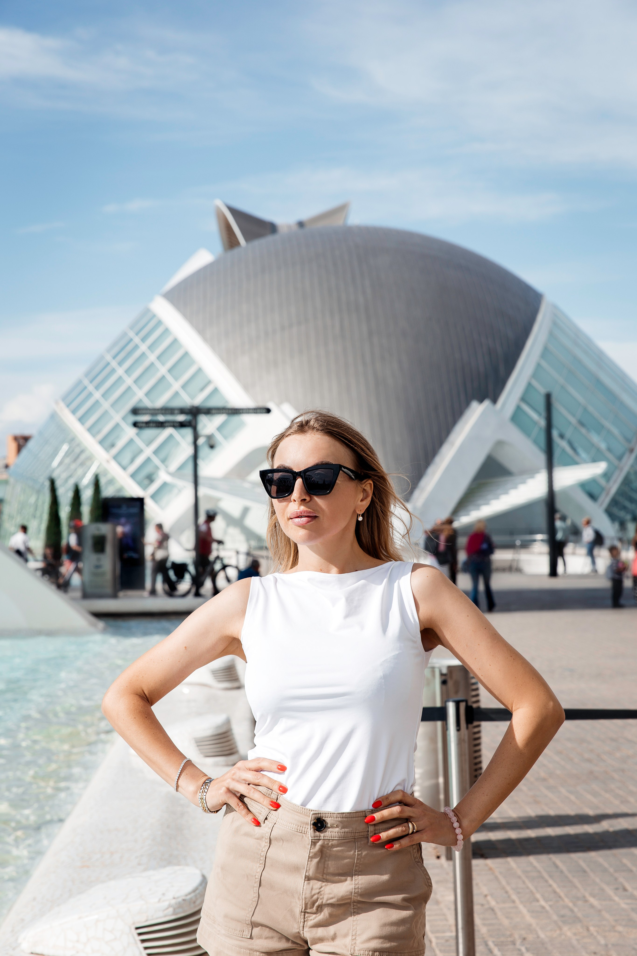 Уверенная в себе женщина в солнцезащитных очках, белом топе и бежевых шортах, позирующая перед знаковым зданием L’Hemisfèric в Городе искусств и наук в Валенсии, Испания