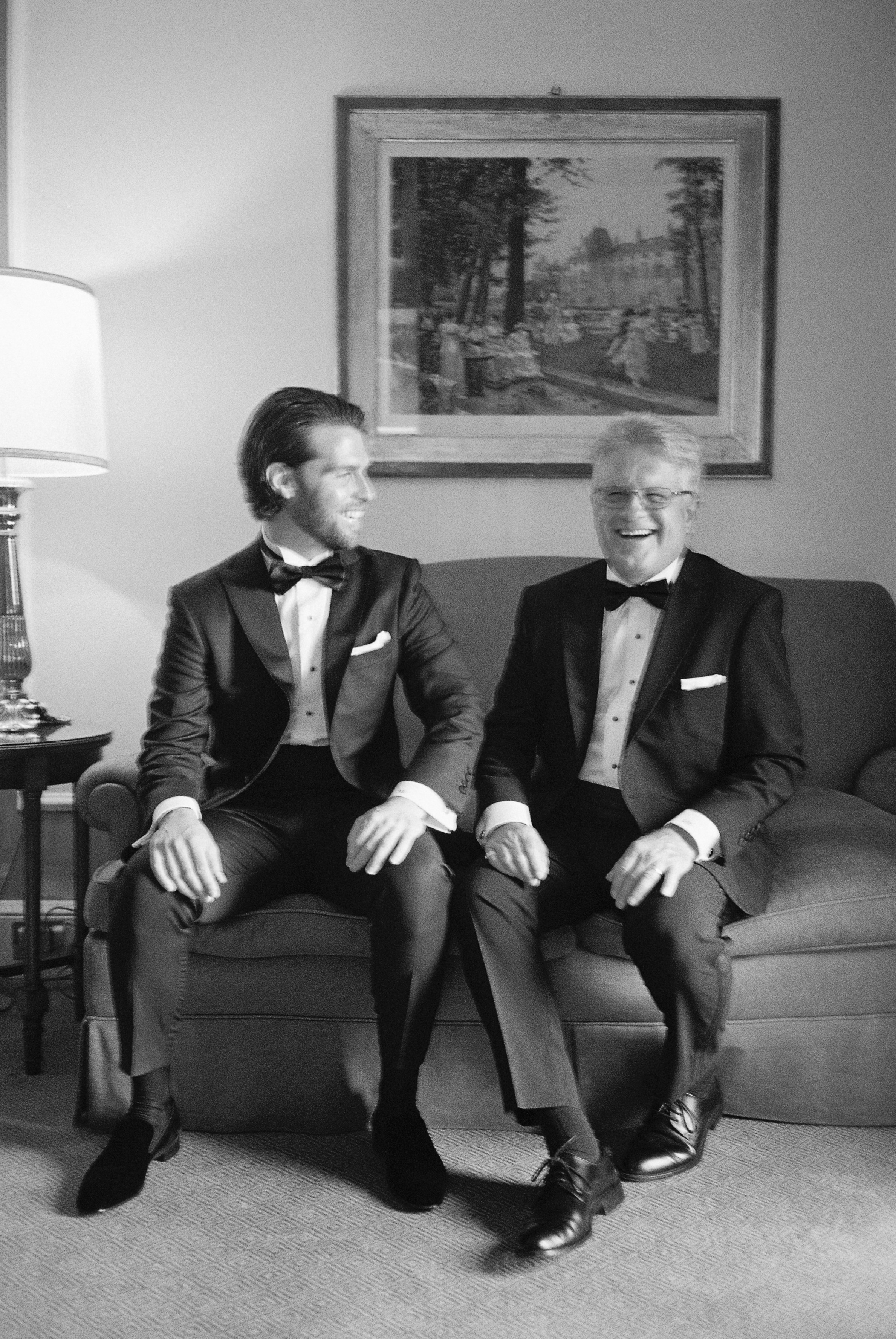 Groom and his father share a joyful moment, seated together in matching black-tie attire.