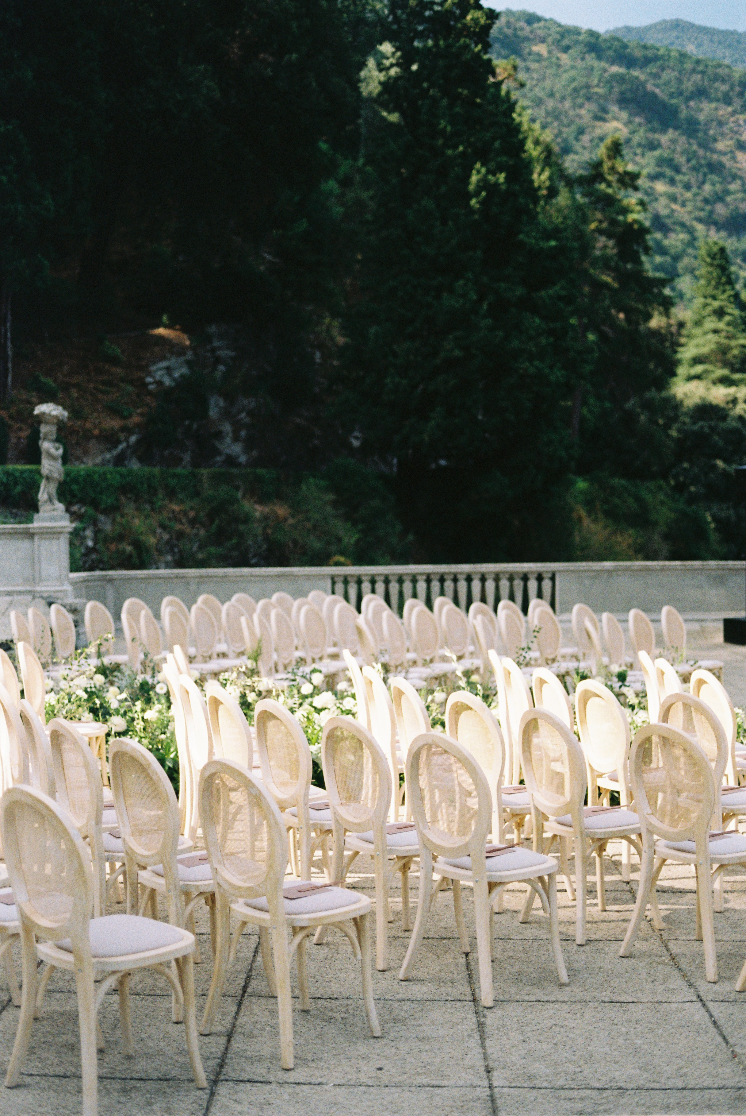 Rows of light-colored chairs set in an outdoor garden, surrounded by lush greenery and mountains.