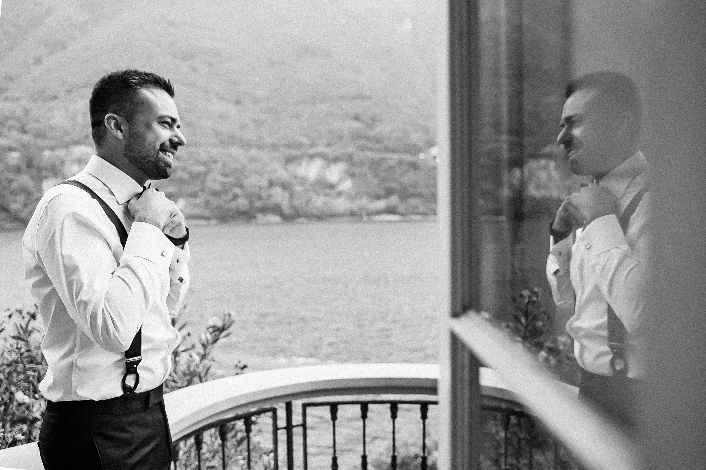 Black and white photo of a groom adjusting a bow tie while facing a mirror, dressed in a white shirt, suspenders, and dark trousers. A scenic lake and mountain view appears in the background, adding a refined, cinematic atmosphere.