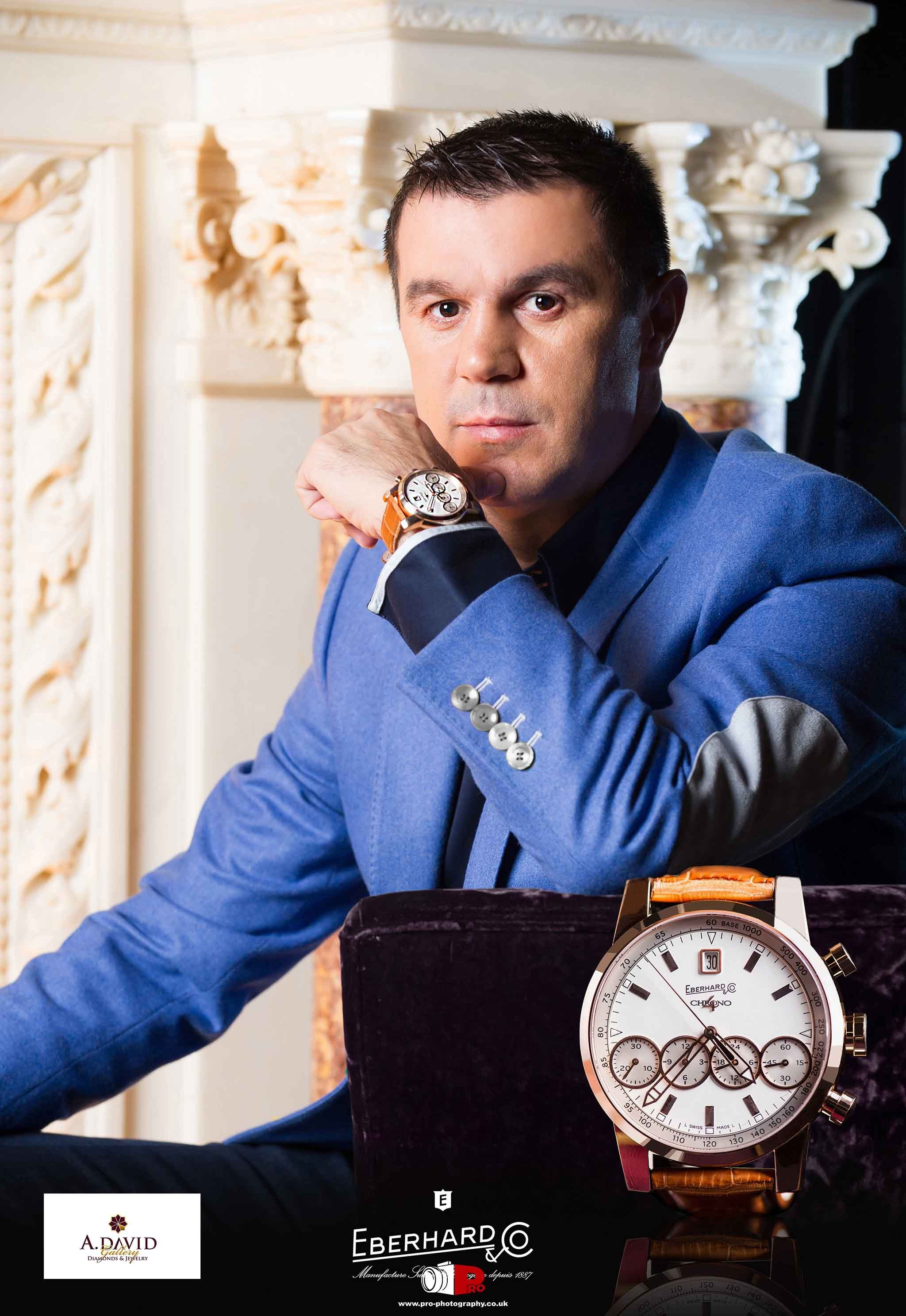 Man in a tailored blue suit showcasing an Eberhard & Co. luxury wristwatch with a leather strap in an elegant indoor setting.