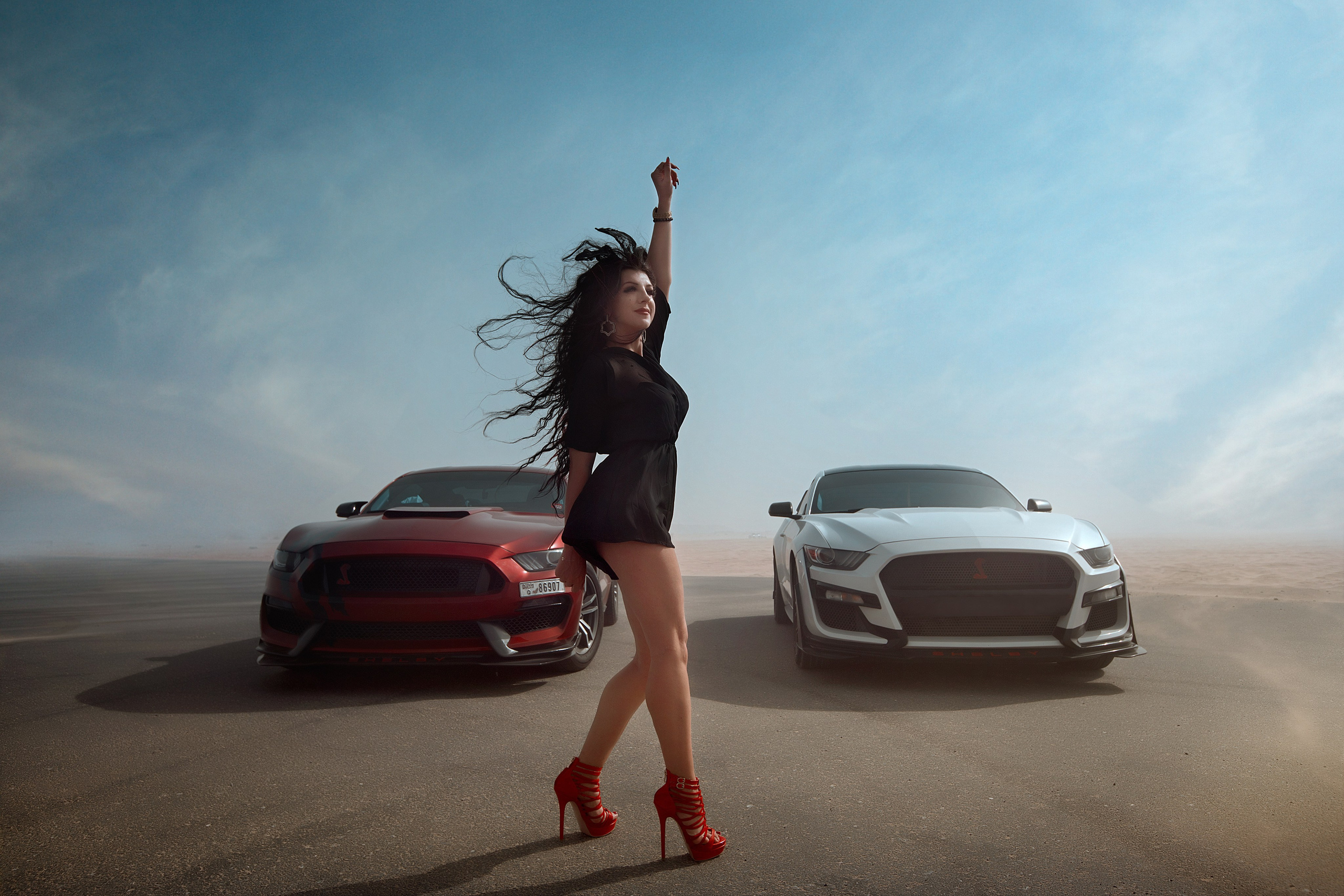 A beautiful model in a luxurious black suit raises her hand against the backdrop of a white exclusive, stunning car, with a red sports car parked nearby, set against the Dubai sandy road