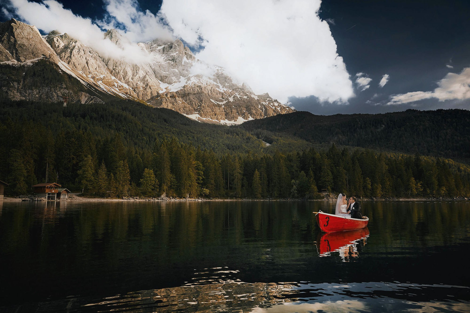Bride and groom in a red rowboat on Lake Eibsee as the Alps rise majestically above them.