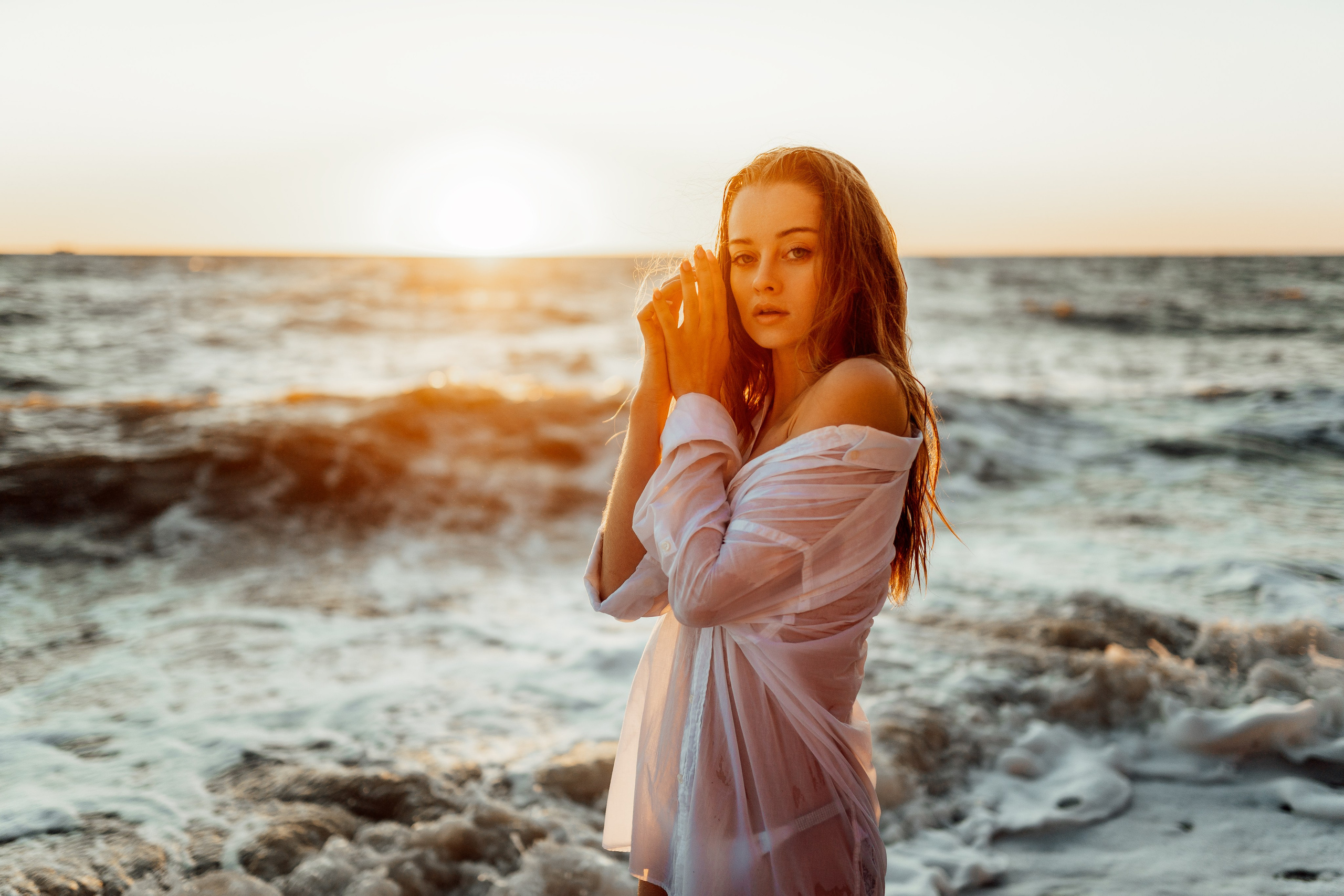 At golden hour, the model stands by the ocean in a white bikini and sheer cover-up, capturing the tranquility of the moment. The warm light and the gentle waves highlight the serene beauty of the scene. Golden hour, tranquility, warm light, gentle waves, serene beauty
