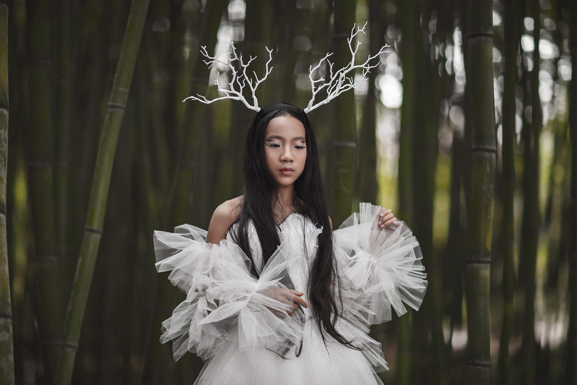 Charming young Chinese girl in a white airy dress, in a bamboo grove. Her head is adorned with graceful white horns. Art photography in Shanghai