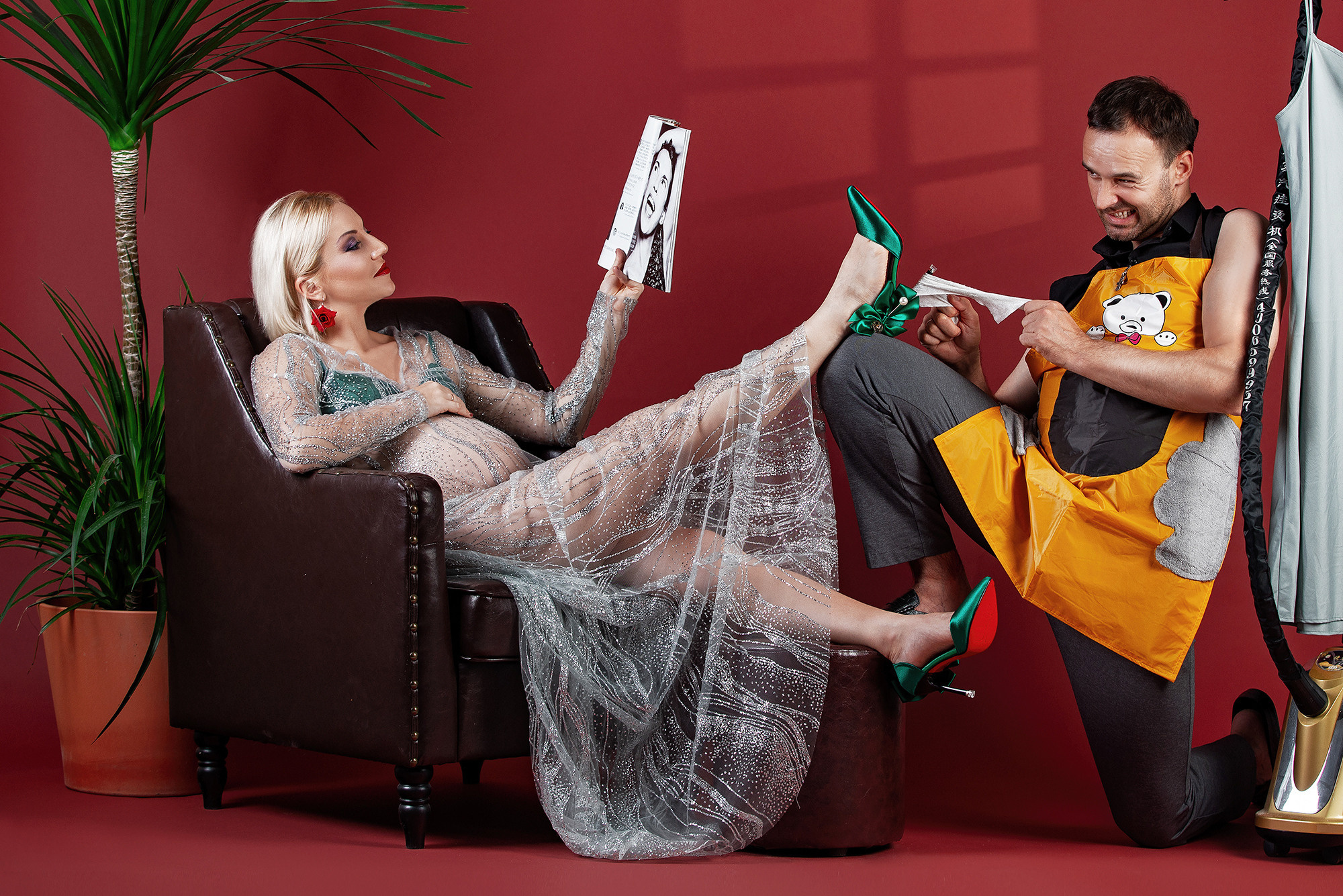 Studio photo shoot in humorous style. Red background, palm tree. Man-houseman shining shoes for his woman.