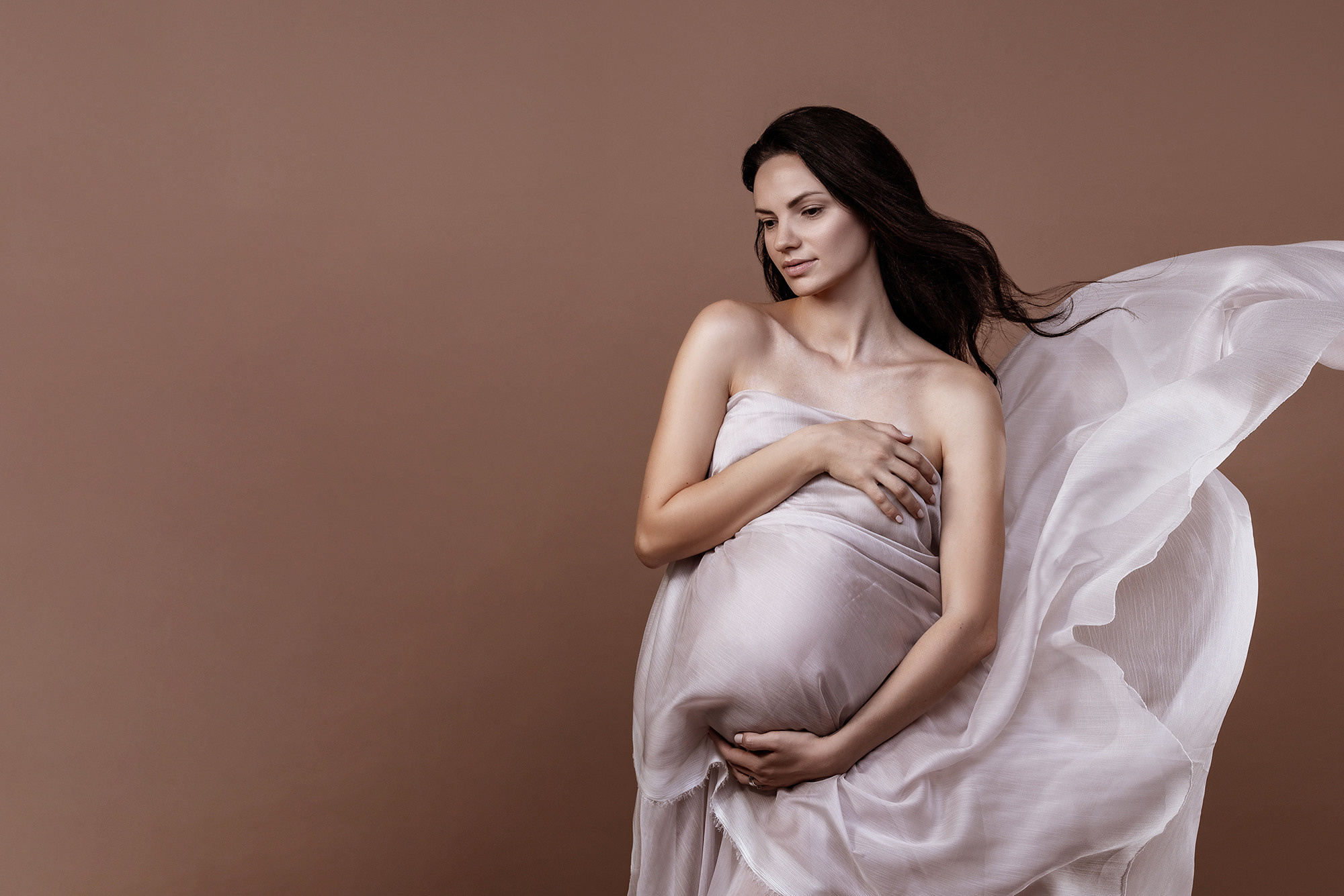 Studio photo shoot for pregnant beautiful woman in flying pink fabric on beige background
