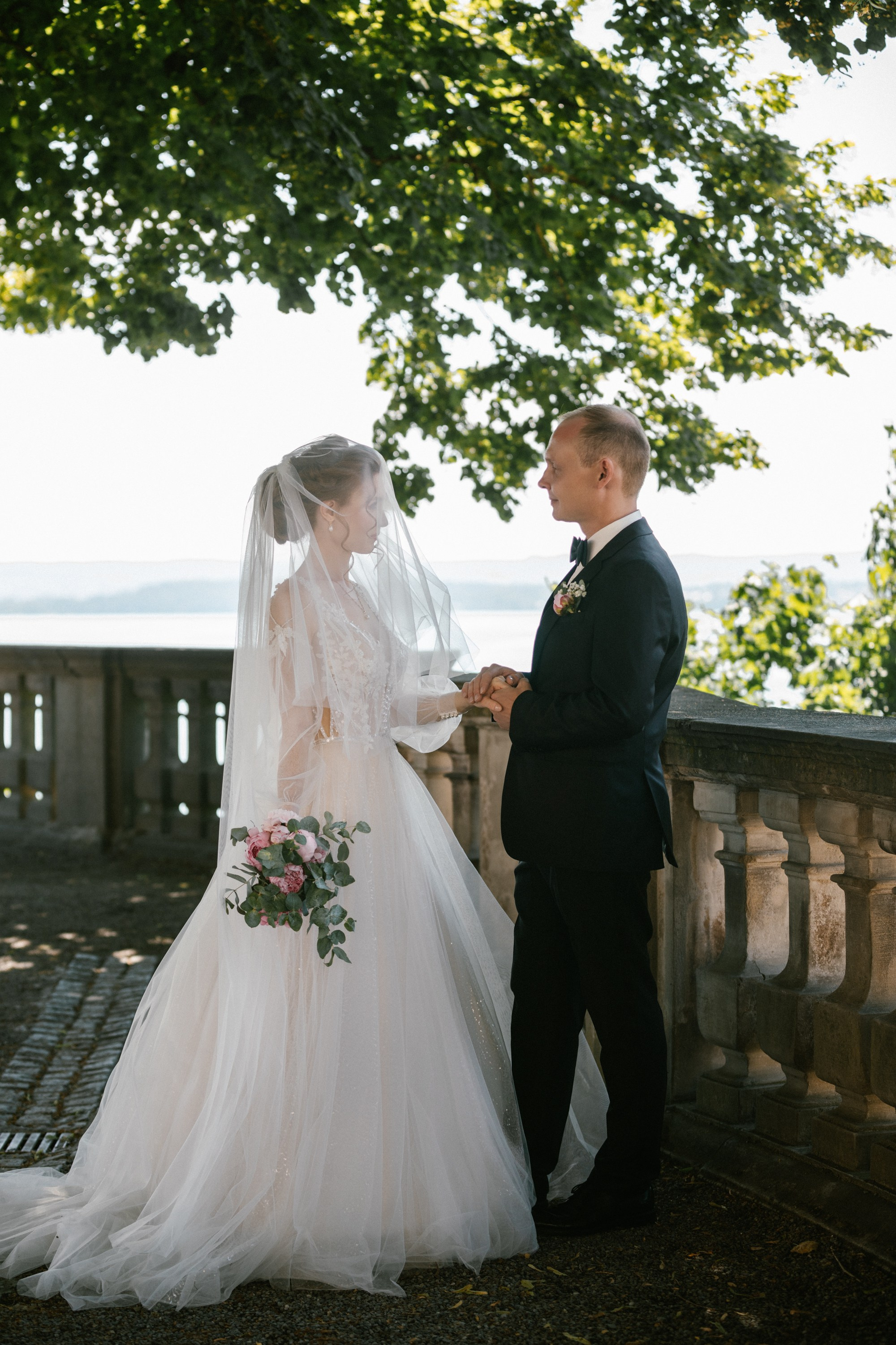 Bride and groom holding hands under tree canopy on lakeview terrace at Schloss Meersburg