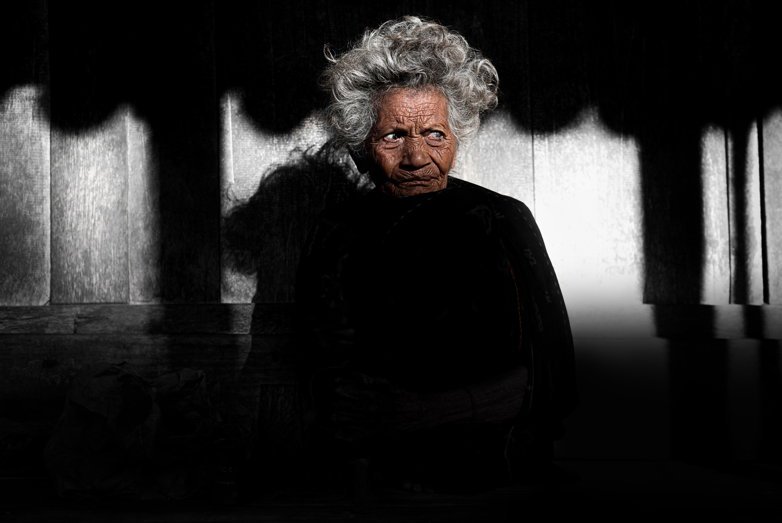 A powerful portrait of an elderly woman sitting in dramatic light and shadow, her textured face and silver hair illuminated against a dark wooden background. The intensity of her gaze and the contrast of light evoke a sense of resilience and untold stories. This image showcases the photographer’s skill in capturing raw emotion and detail, making it ideal for clients seeking evocative visual storytelling for editorial or documentary projects.