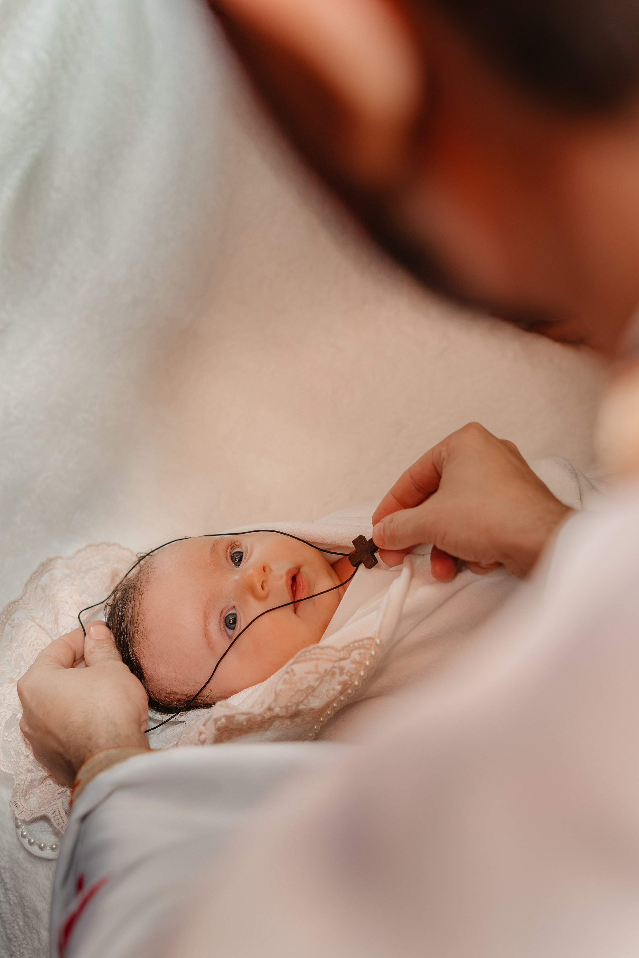 Preotul pune o cruciuliță sfântă la gâtul pruncului Eveline Maria, după scăldătoarea botezului. Fetița este înfășată în prosoape albe curate. Un moment de binecuvântare și încheiere a tainei.