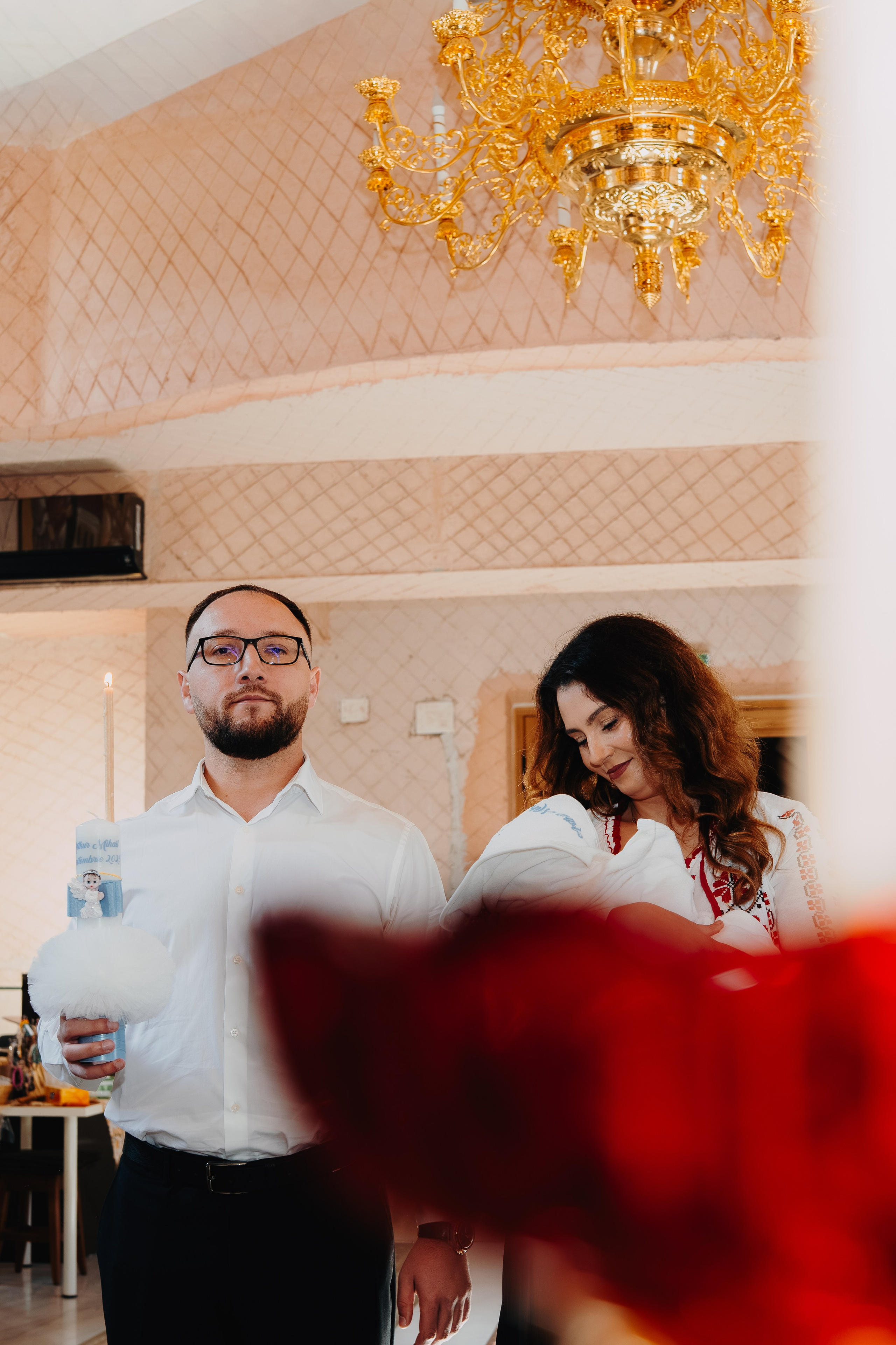 Fotografie în culori calde de după botezul lui Arthur Mihail în Catedrala Bârlad. Flacăra portocalie a lumânării de botez, ținută de naș, luminează scenea intimă în care nașa îl privește pe prunc cu dragoste. Detaliile cromatică ale catedralei sunt vizibile.