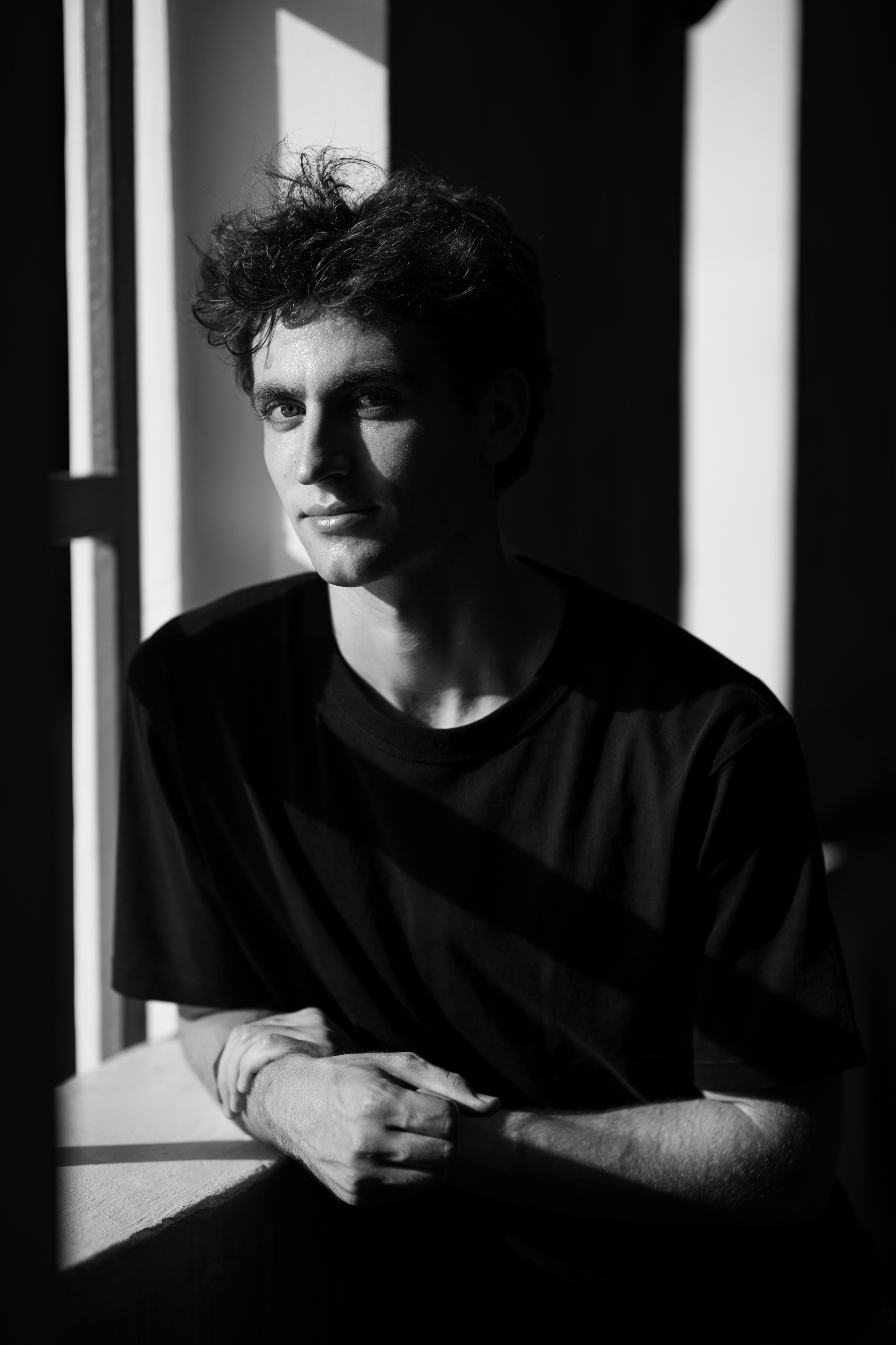 Monochrome portrait in window light—subject leaning on sill with crossed hands; bold diagonal shadow pattern, photographed in Singapore.