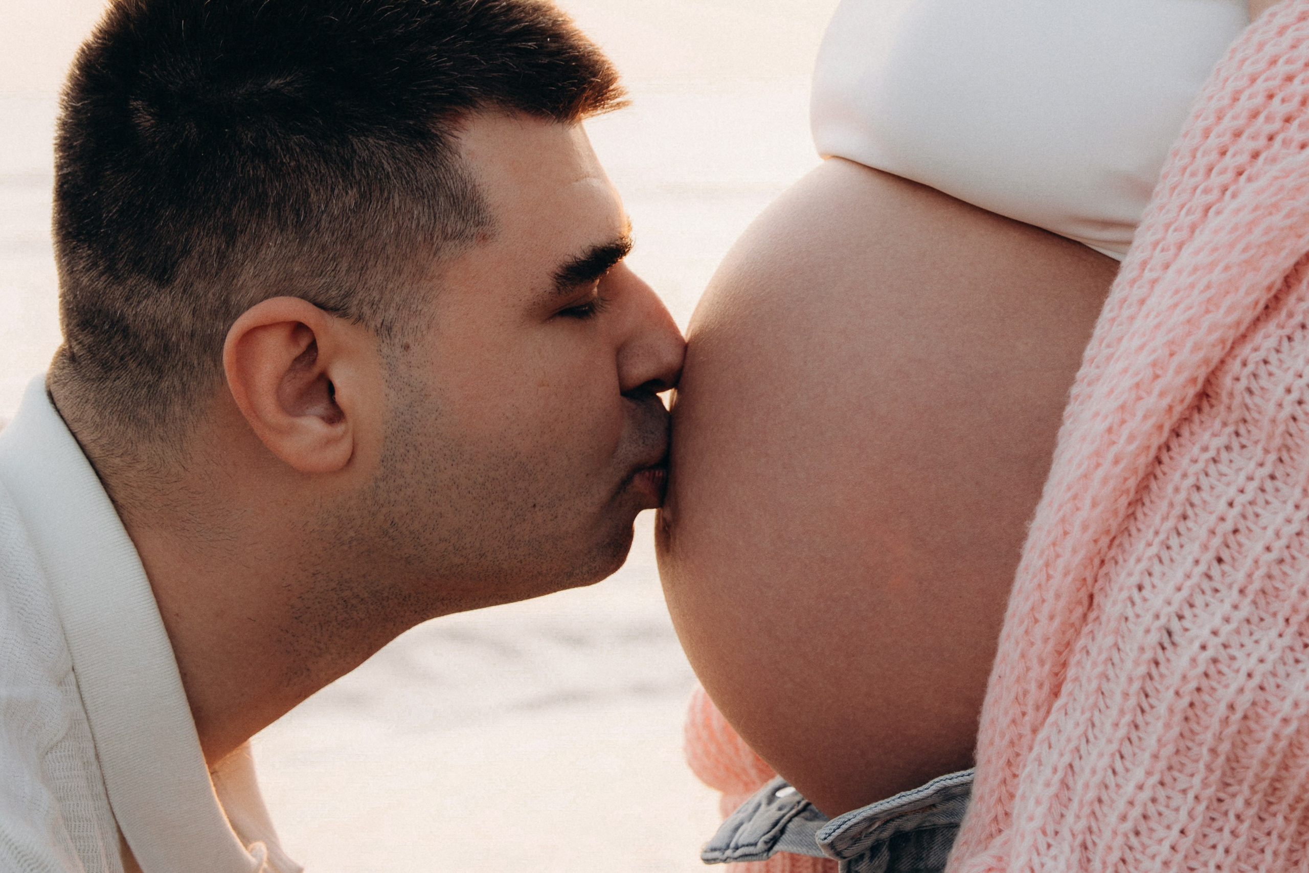 Sesión de fotos de embarazo en Valencia, España, con un padre besando la barriga de embarazada junto al mar, capturada con luz natural suave para un momento íntimo y emocional — ideal para sesiones maternity en Valencia y en toda España.