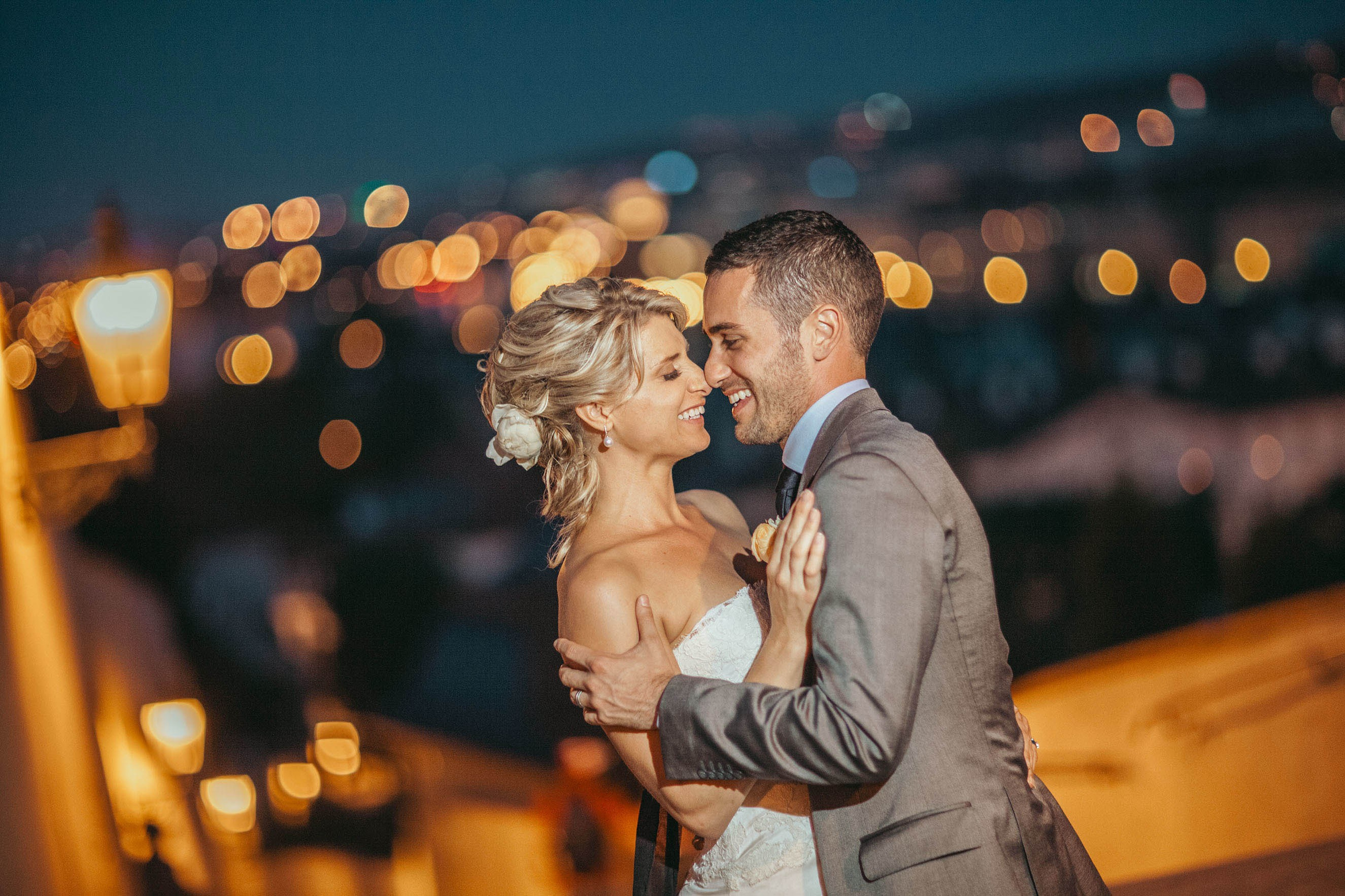 A beaming bride and groom snuggle up as they laugh hysterically against the background of twinkling city lights overlooking Prague at the Villa Richter.