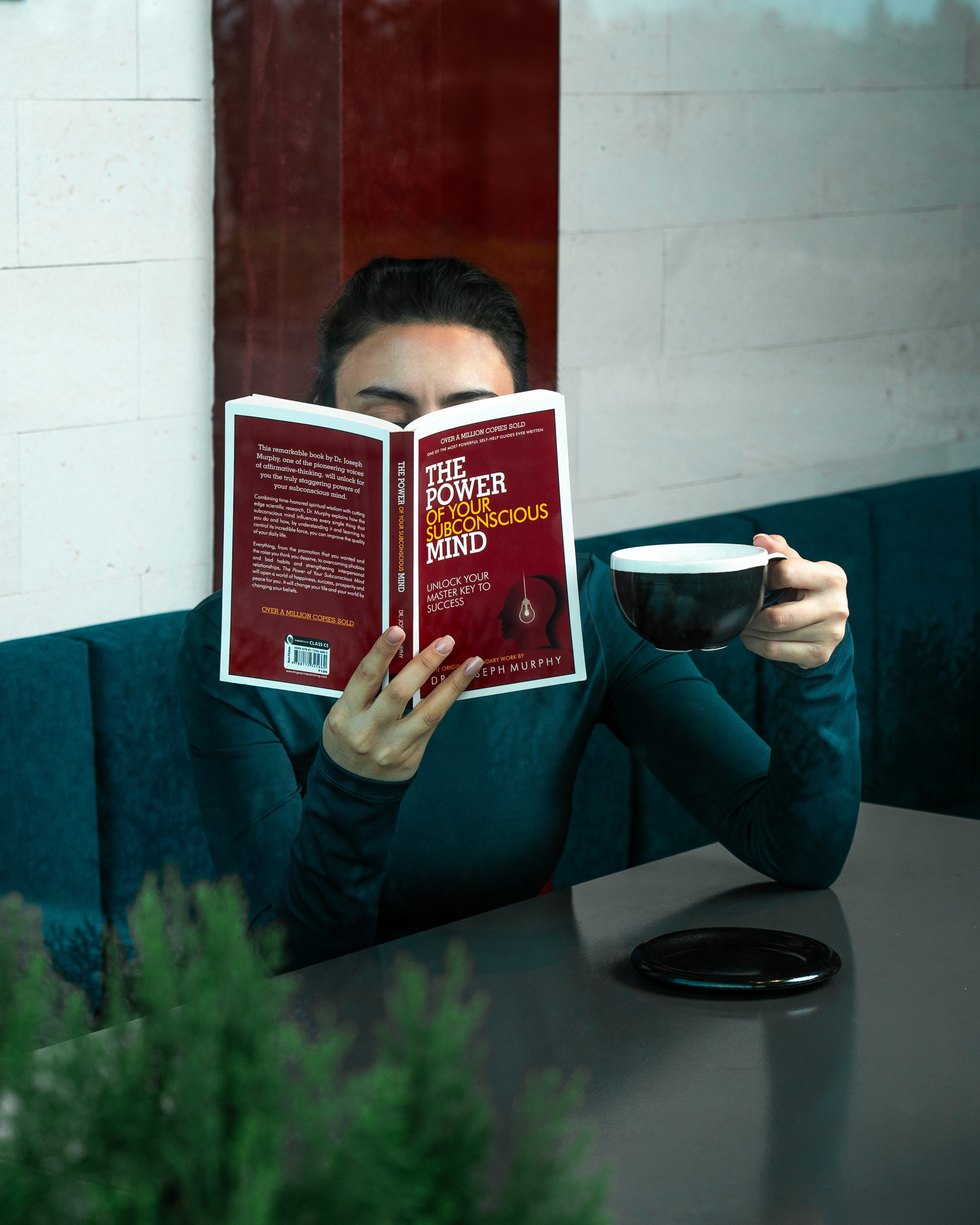 Kafedə əlində qara qəhvə fincanı tutarkən Cozef Murfinin "Şüuraltı Ağlınızın Gücü"nü oxuyan şəxs.