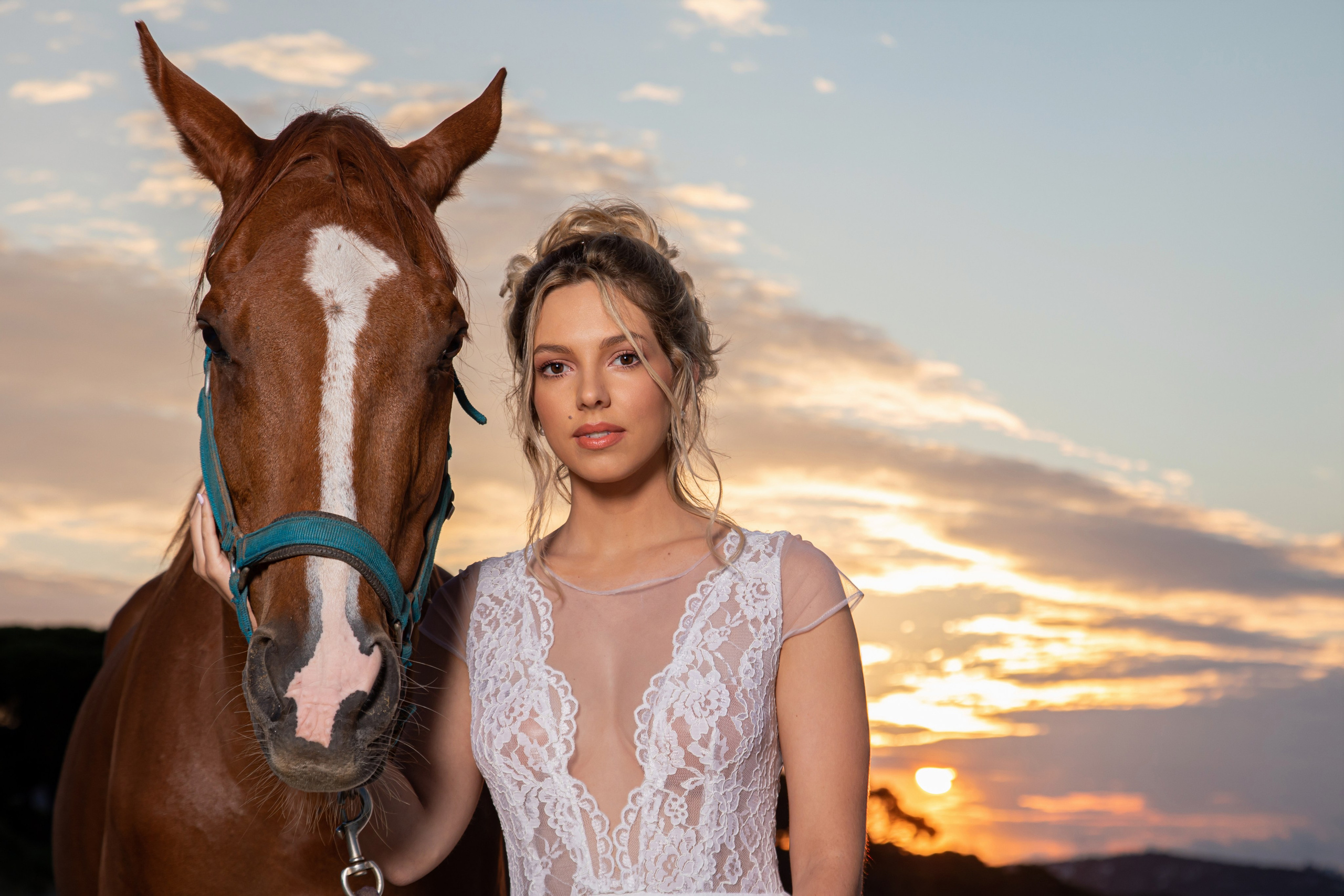 A young woman with blonde hair in an elegant white lace dress and a brown horse with a white blaze stand together on a hill at sunset, silhouetted by the warm light and clouds in the sky.
