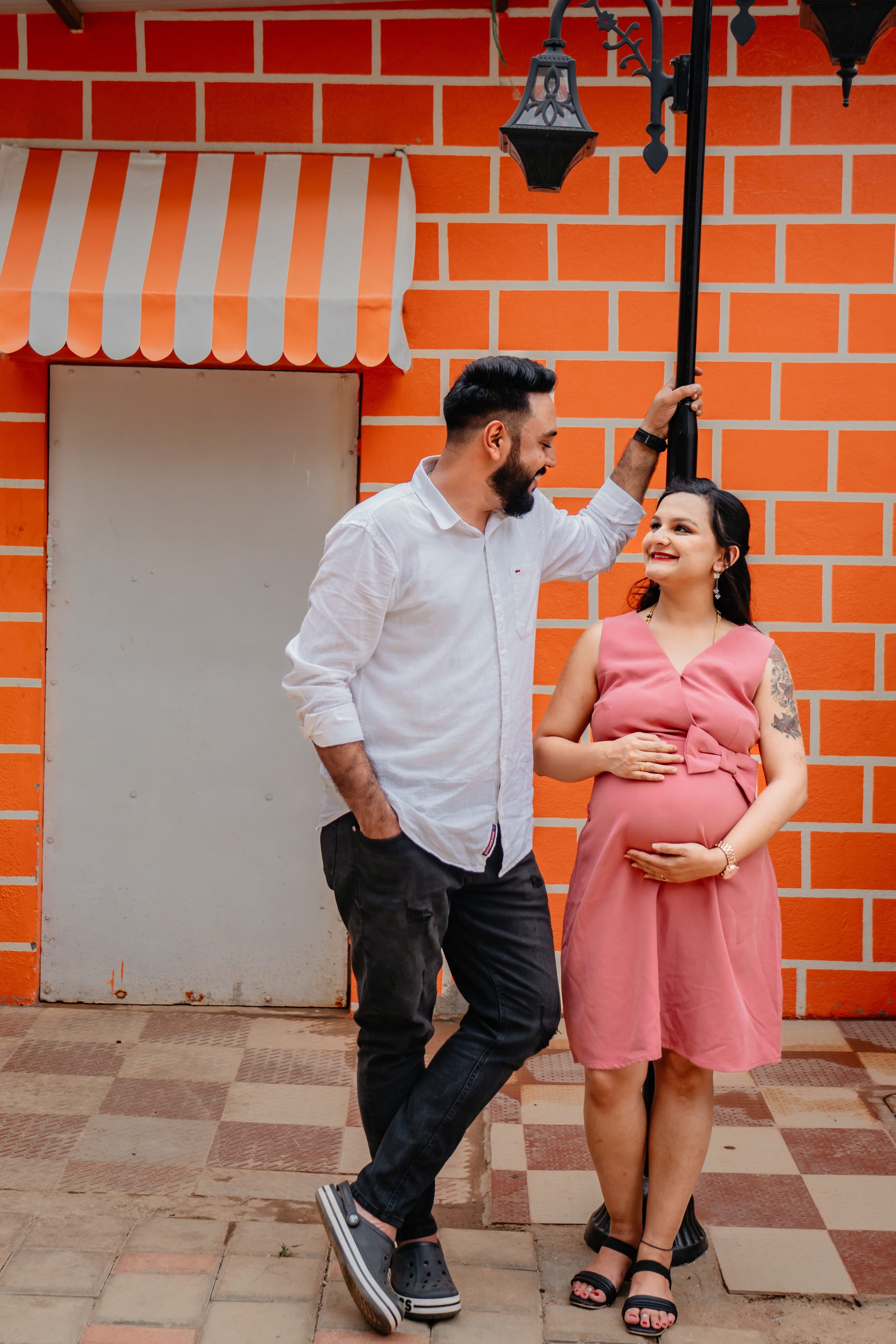 Outdoor maternity photoshoot in Bengaluru featuring a man in a white shirt and a woman in a pink dress posing against a vibrant orange brick wall backdrop with a street lamp.