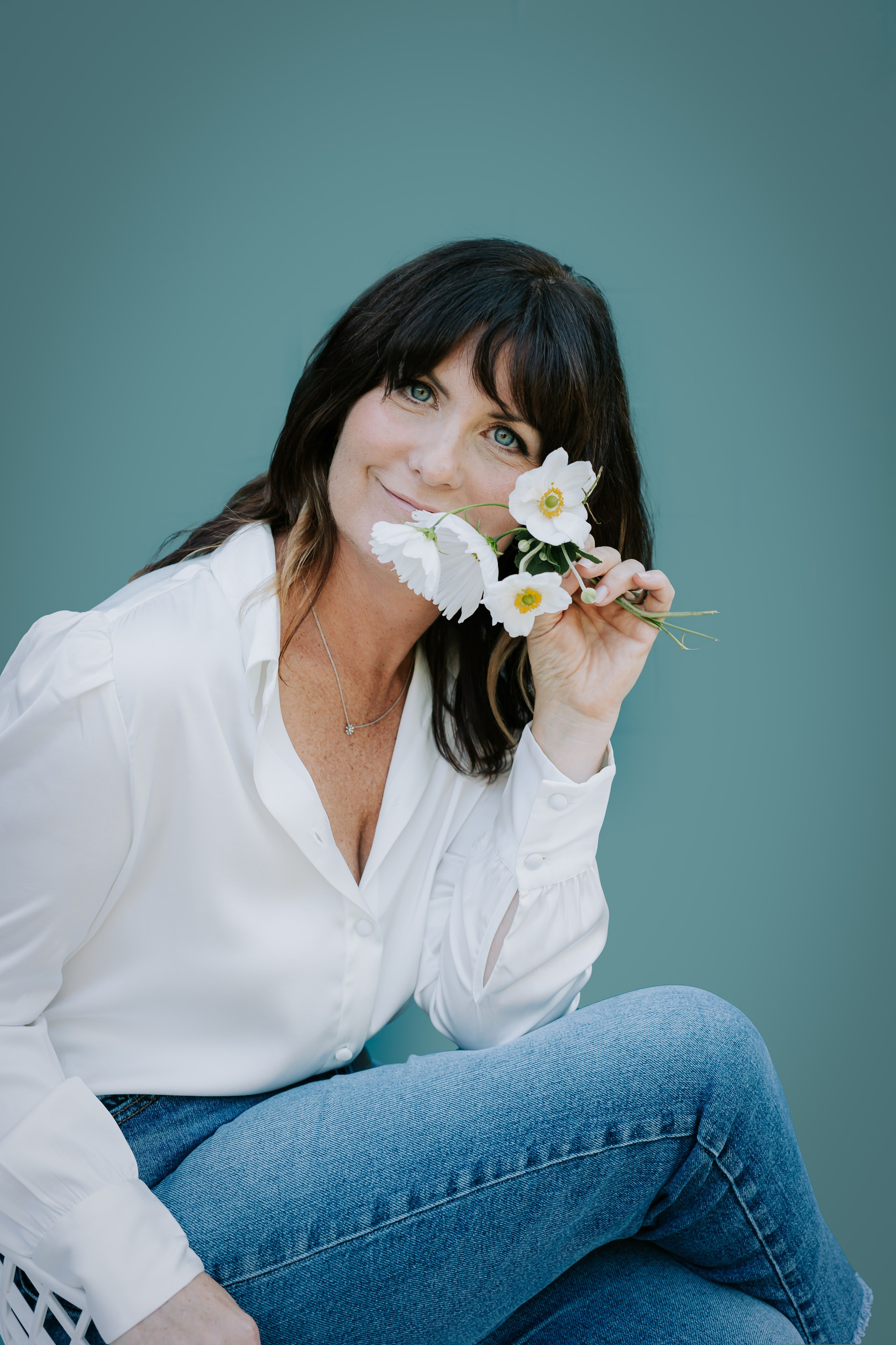 headshot of woman in white top, holding white flower in front of her eye on a teal backdrop 