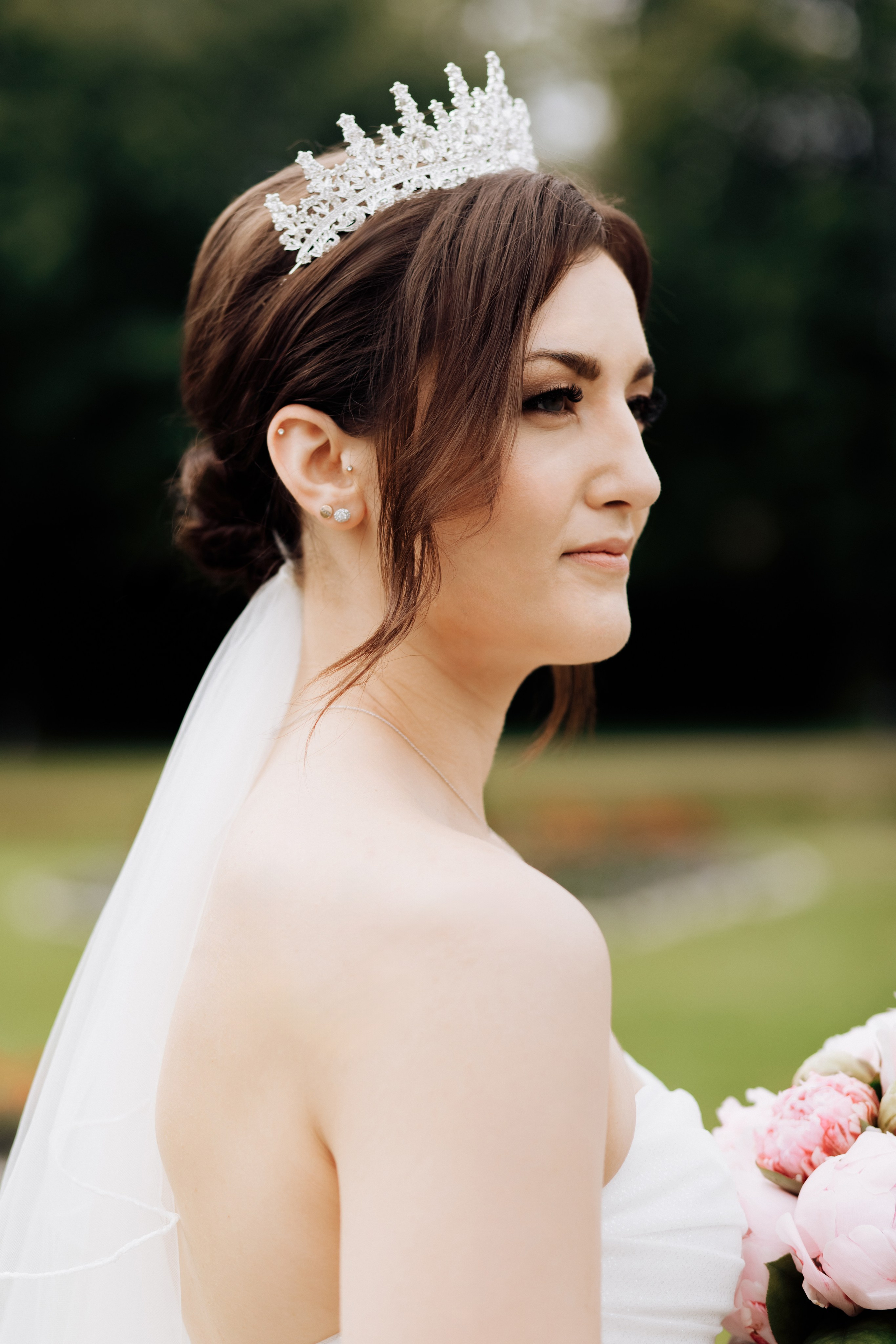 Die Braut schaut stolz in die Ferne, trägt eine Krone und ein elegantes Hochzeitskleid mit offenen Schultern. Ein majestätischer Moment der Hochzeit.