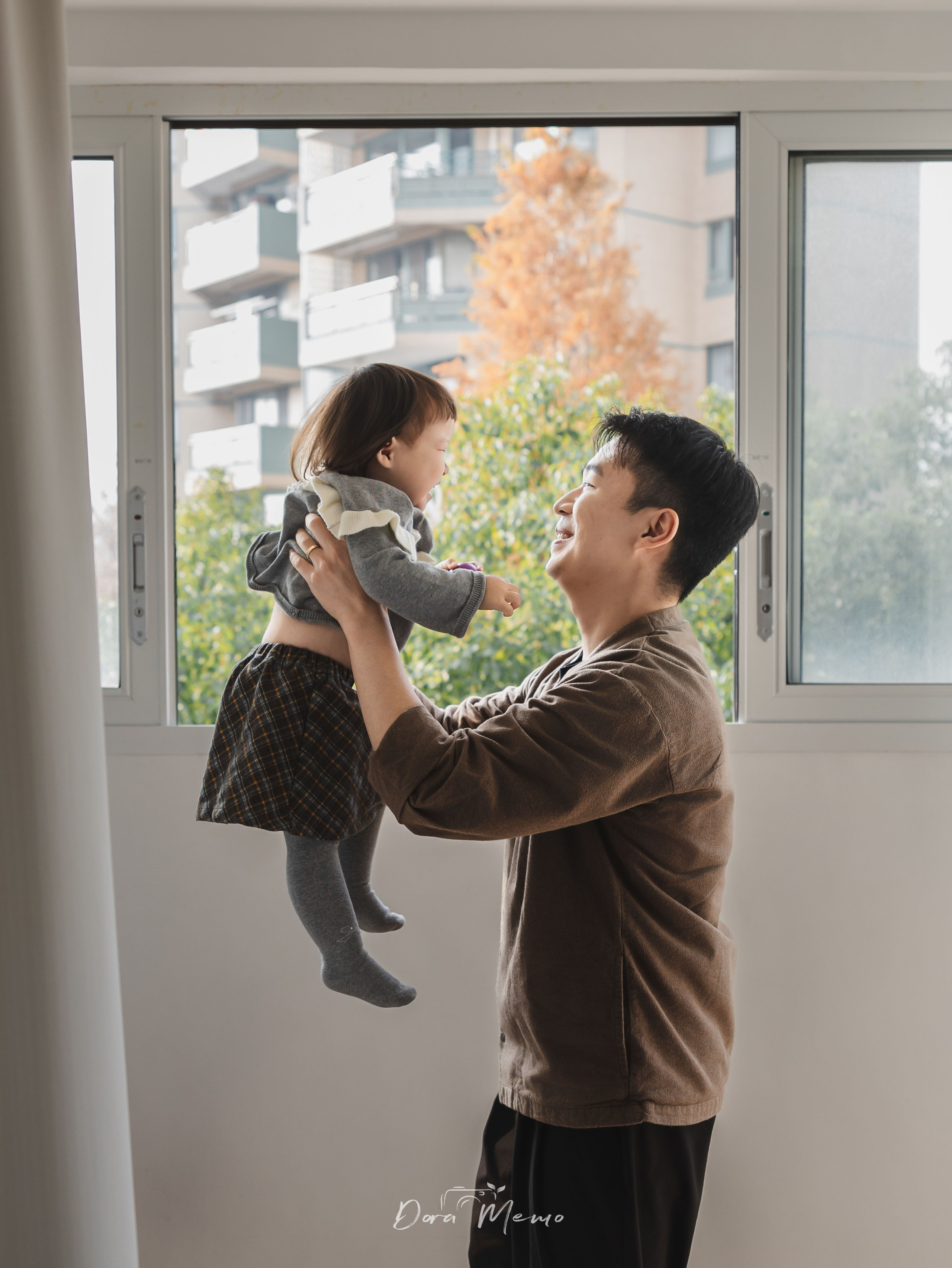 Father lifting his happy toddler daughter in the air near a bright window, documentary family photography in Shanghai capturing pure joy.