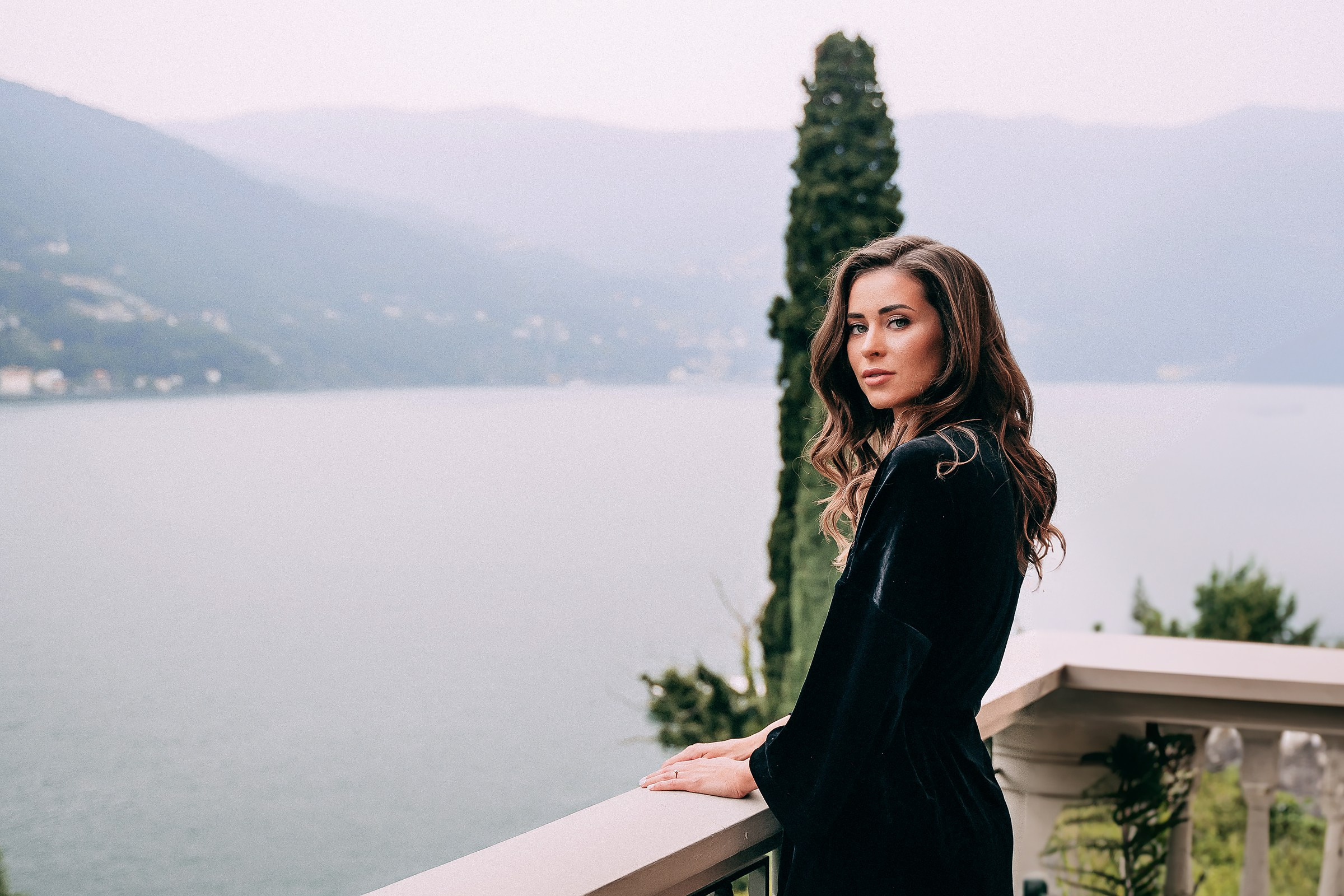 The bride with long, wavy hair stands on a balcony overlooking a tranquil lake encircled by misty mountains. Dressed in a dark outfit, framed by greenery and a tall, slender tree, evoking a serene and introspective mood.