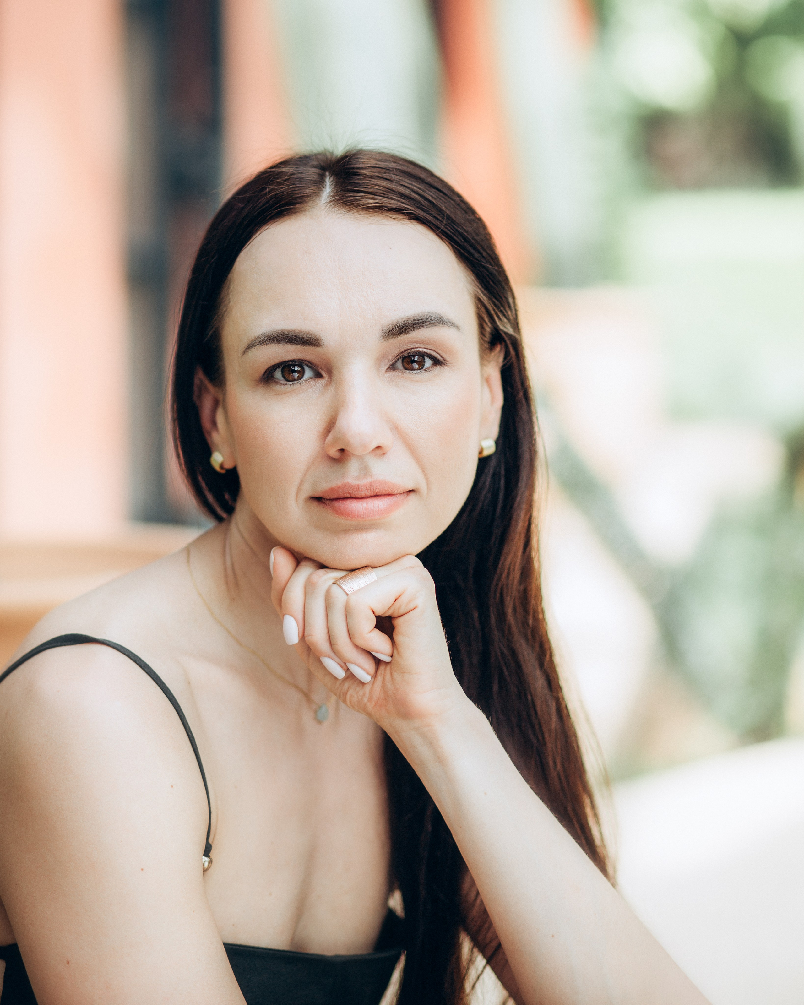 Крупный портрет при естественном освещении женщины с длинными каштановыми волосами, опирающейся подбородком на руку, в черном топе и с мягким макияжем, снят в Кальпе, Испания — интимная и элегантная портретная фотография для тех, кто ищет вневременную фотосессию в Кальпе и Испании.