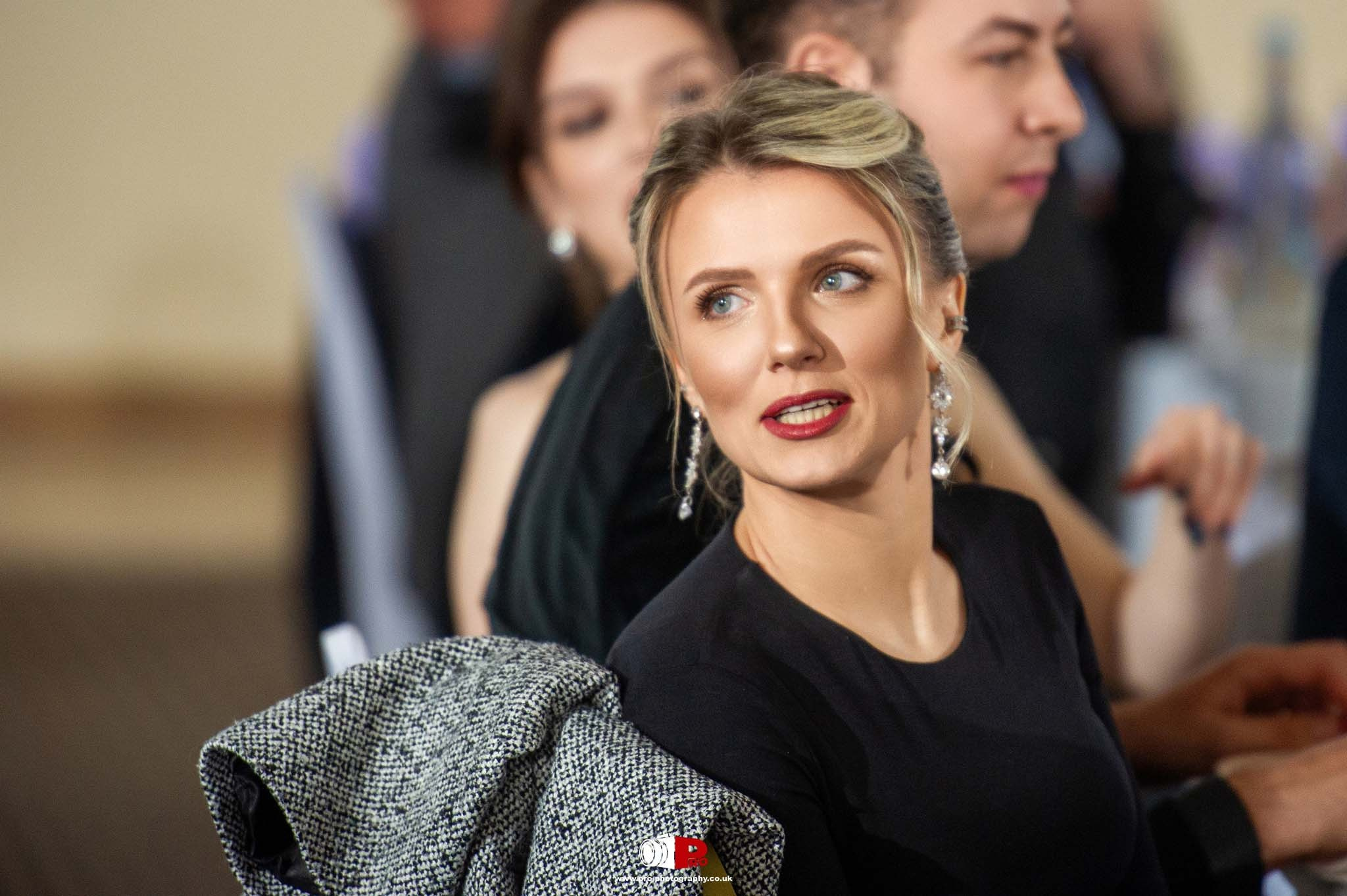 A stylish woman with blonde hair and earrings converses at a formal gathering.