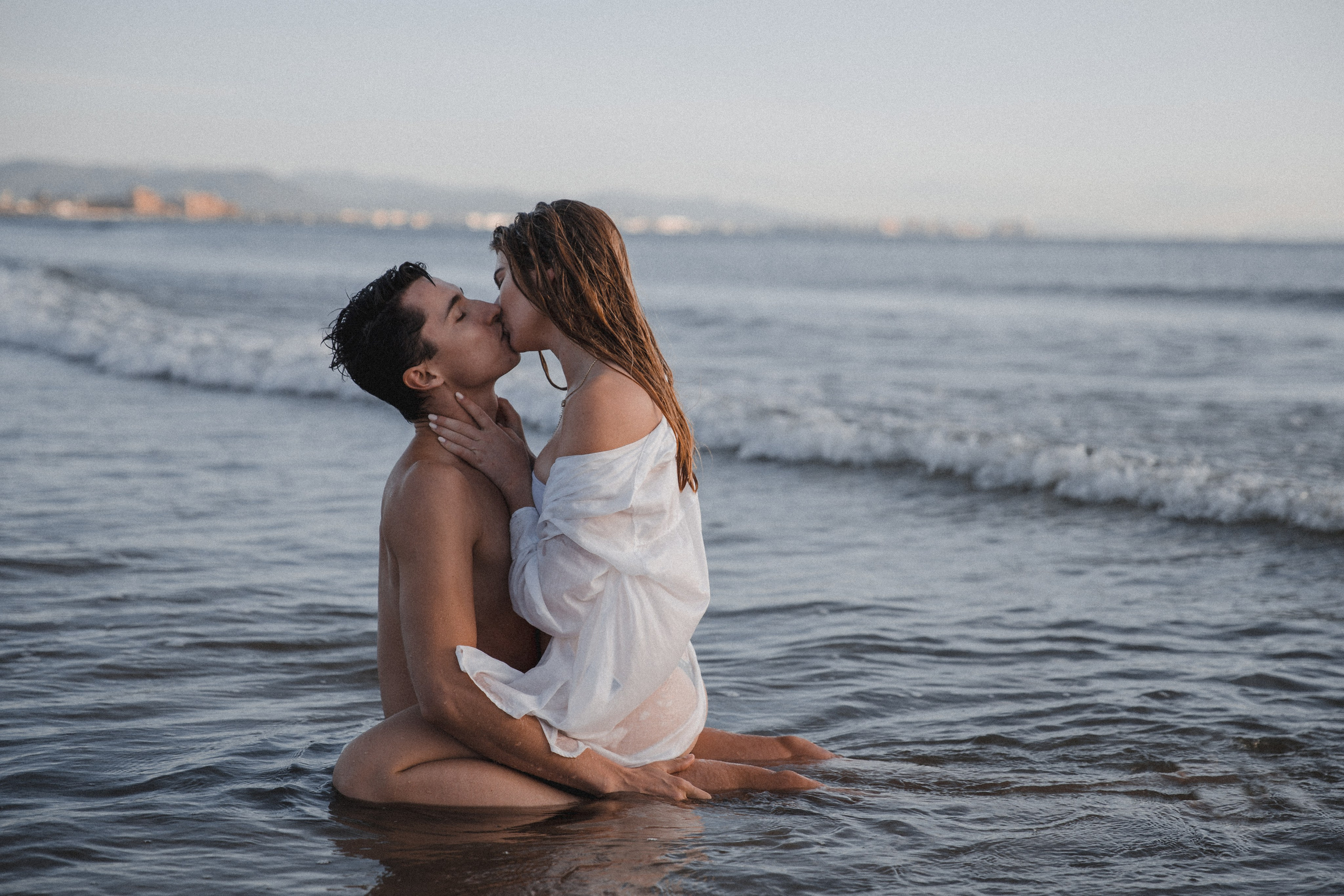 Romantic couple shares a passionate kiss while kneeling in the shallow waves of the Mediterranean Sea during golden hour in Valencia, Spain, with a serene coastline and distant cityscape in the background.