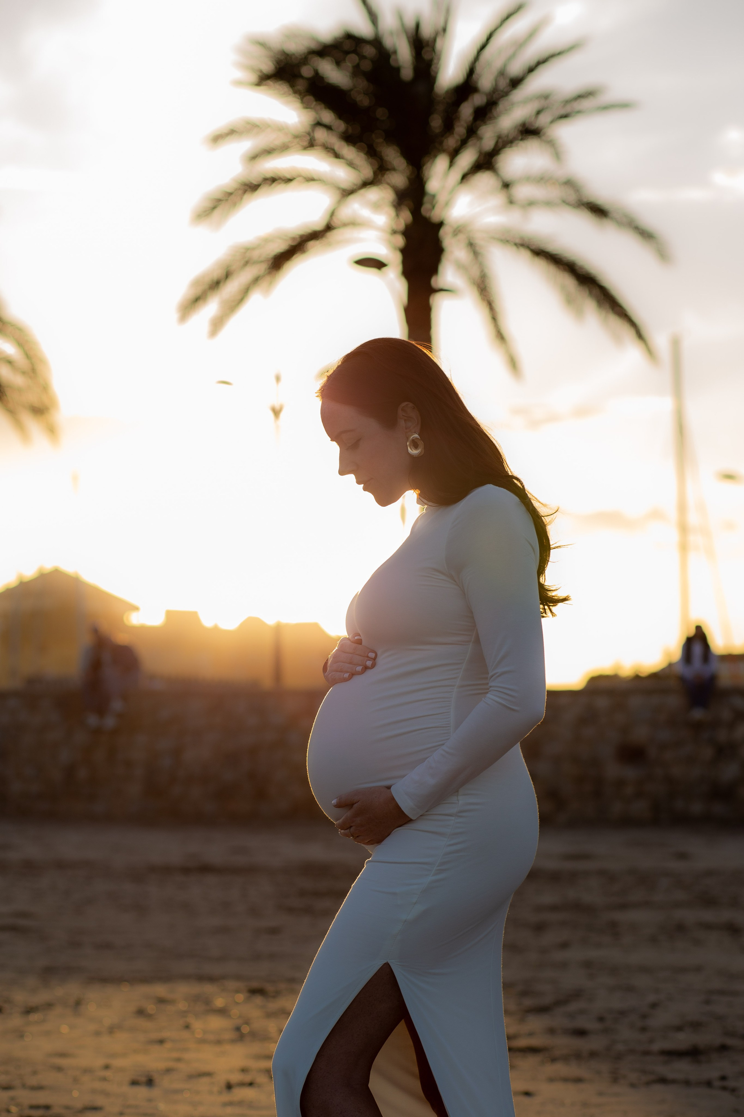 Retrato de maternidad en silueta en Valencia, España — mujer con vestido blanco elegante sosteniendo su vientre durante la hora dorada en la playa, con palmeras y luz suave del atardecer.