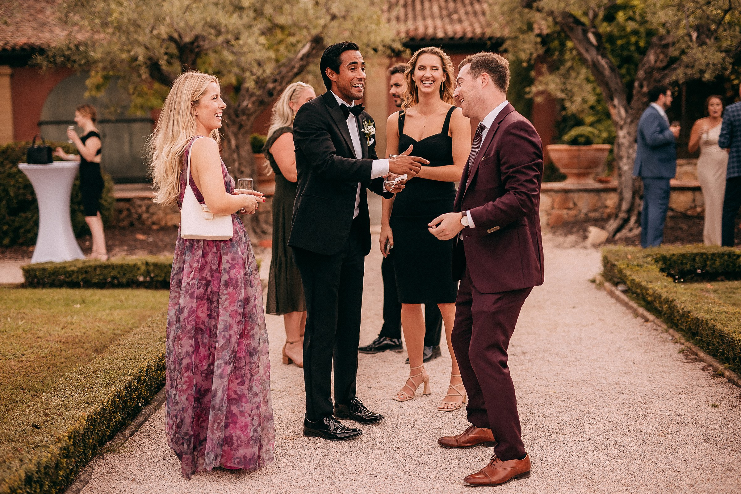 Wedding guests laughing and conversing during an outdoor reception, surrounded by garden pathways and Mediterranean-style architecture.