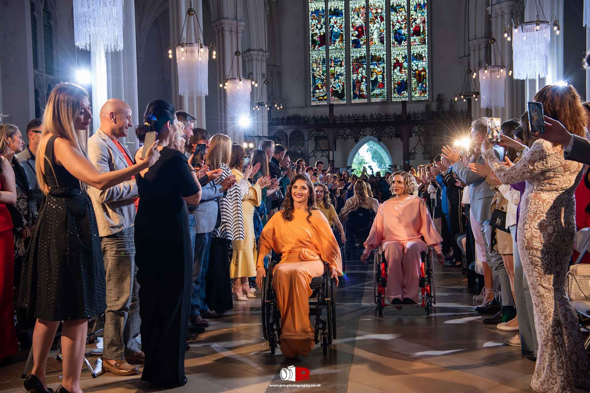 A vibrant fashion show featuring two models in wheelchairs receiving applause in a grand cathedral setting.