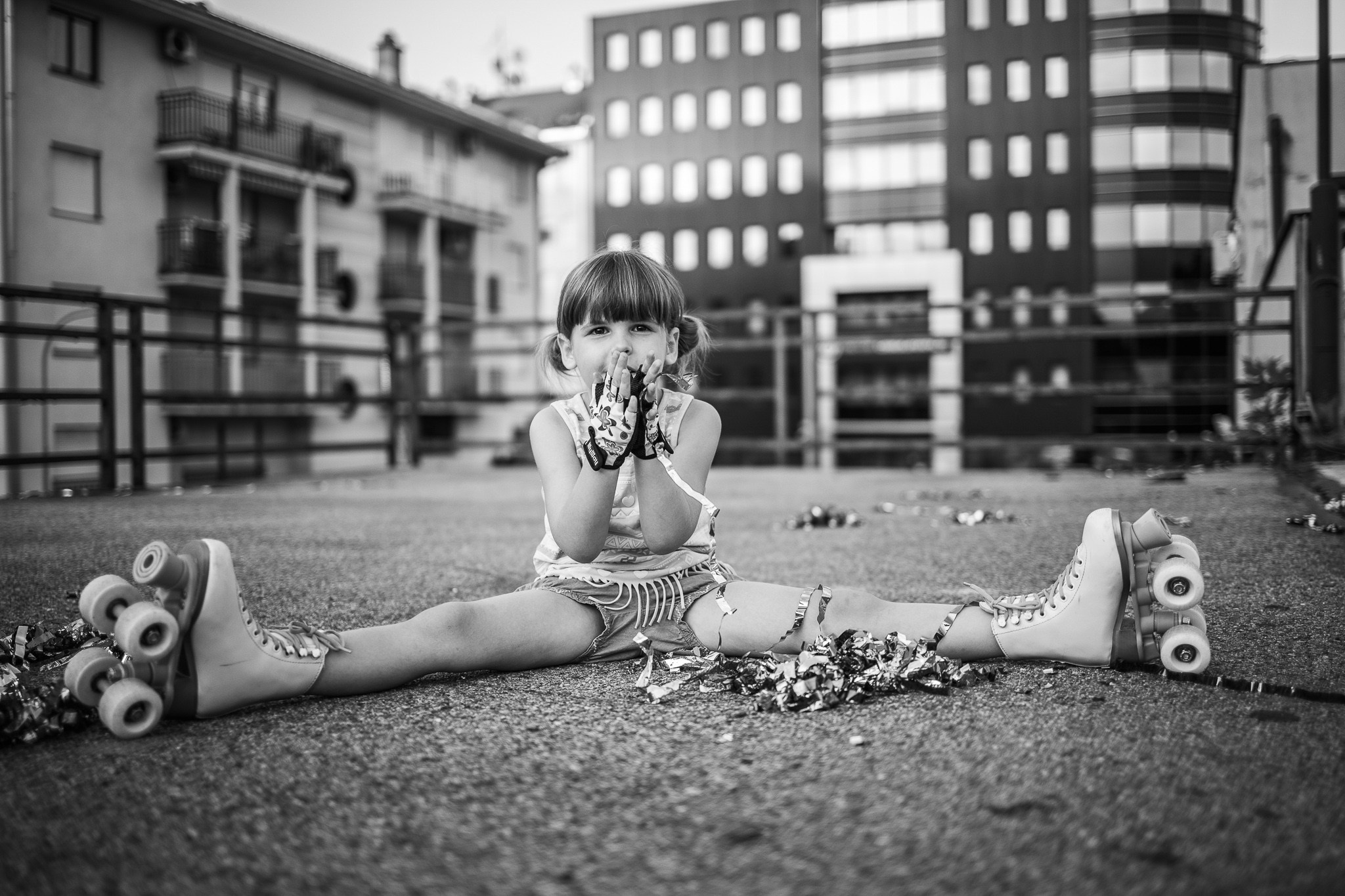 Crno-bela fotografija mlade devojčice sa kikicama na rolerima koja sedi na tlu sa konfeti u rukama, u urbanom okruženju.