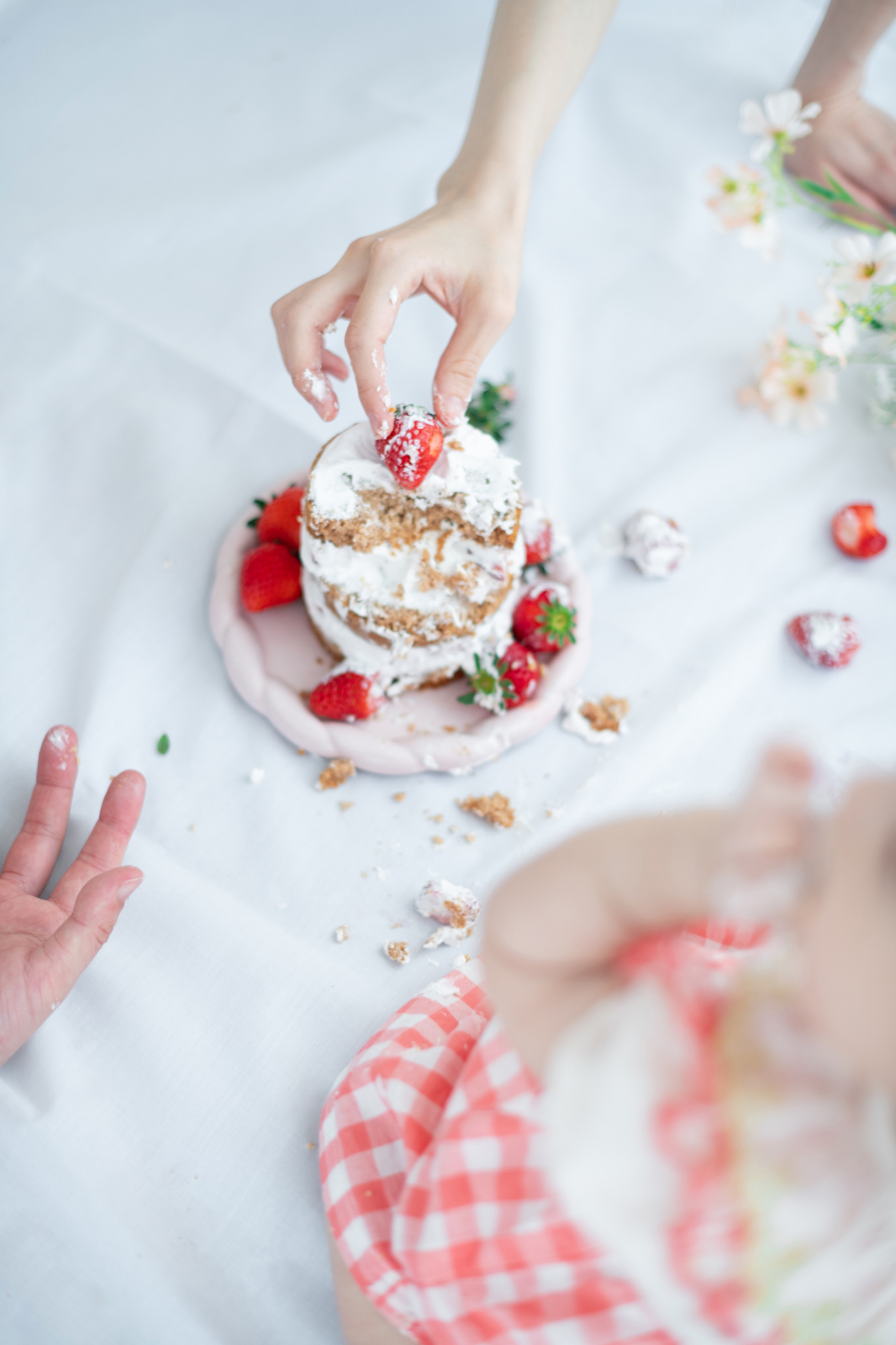 Overhead shot: parent adds a strawberry to a half-smashed mini cake while baby in gingham dress reaches in.