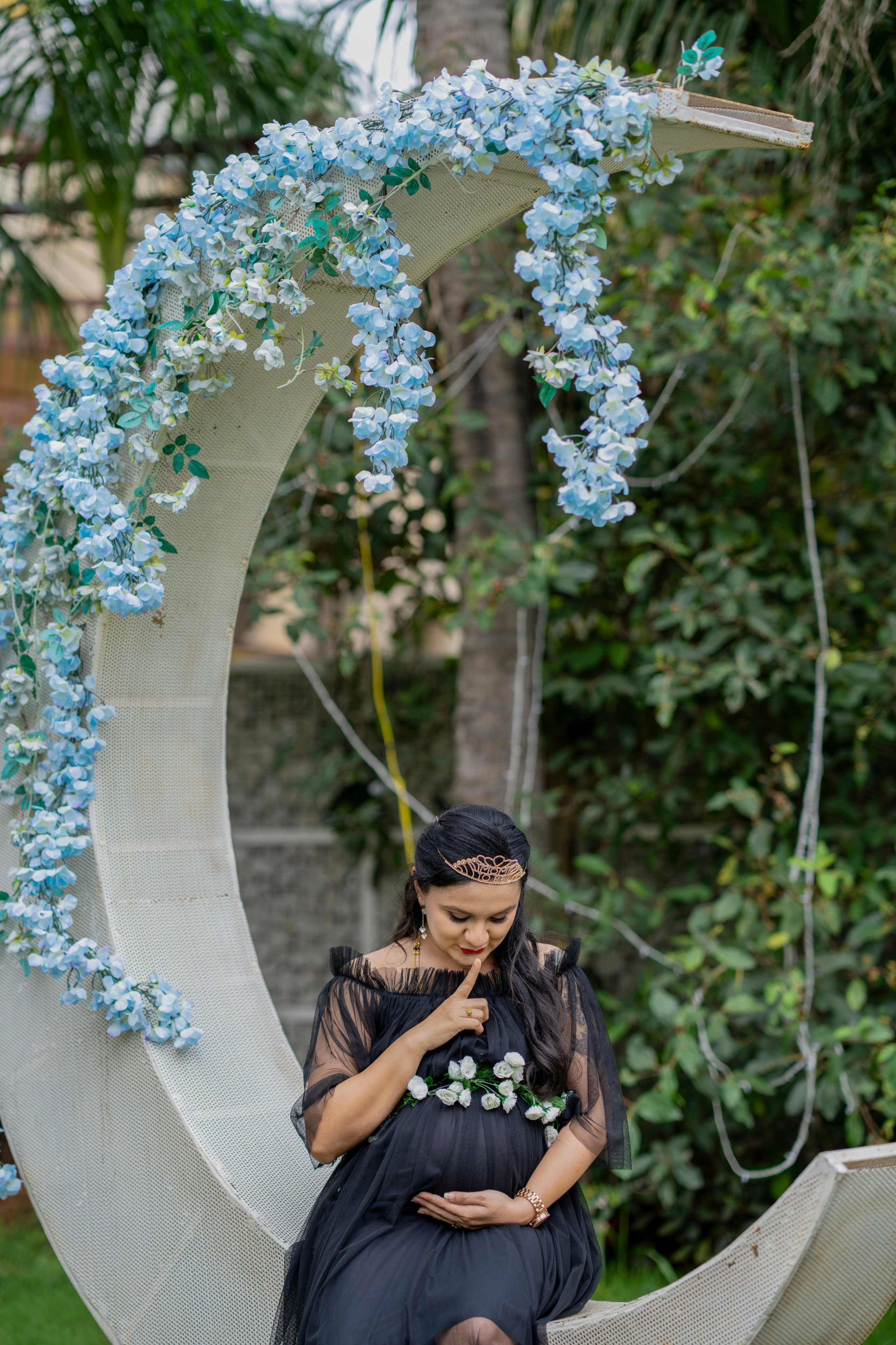 Outdoor maternity photoshoot in Bengaluru featuring a woman in a black tulle gown sitting on a large white crescent moon prop decorated with blue flowers.