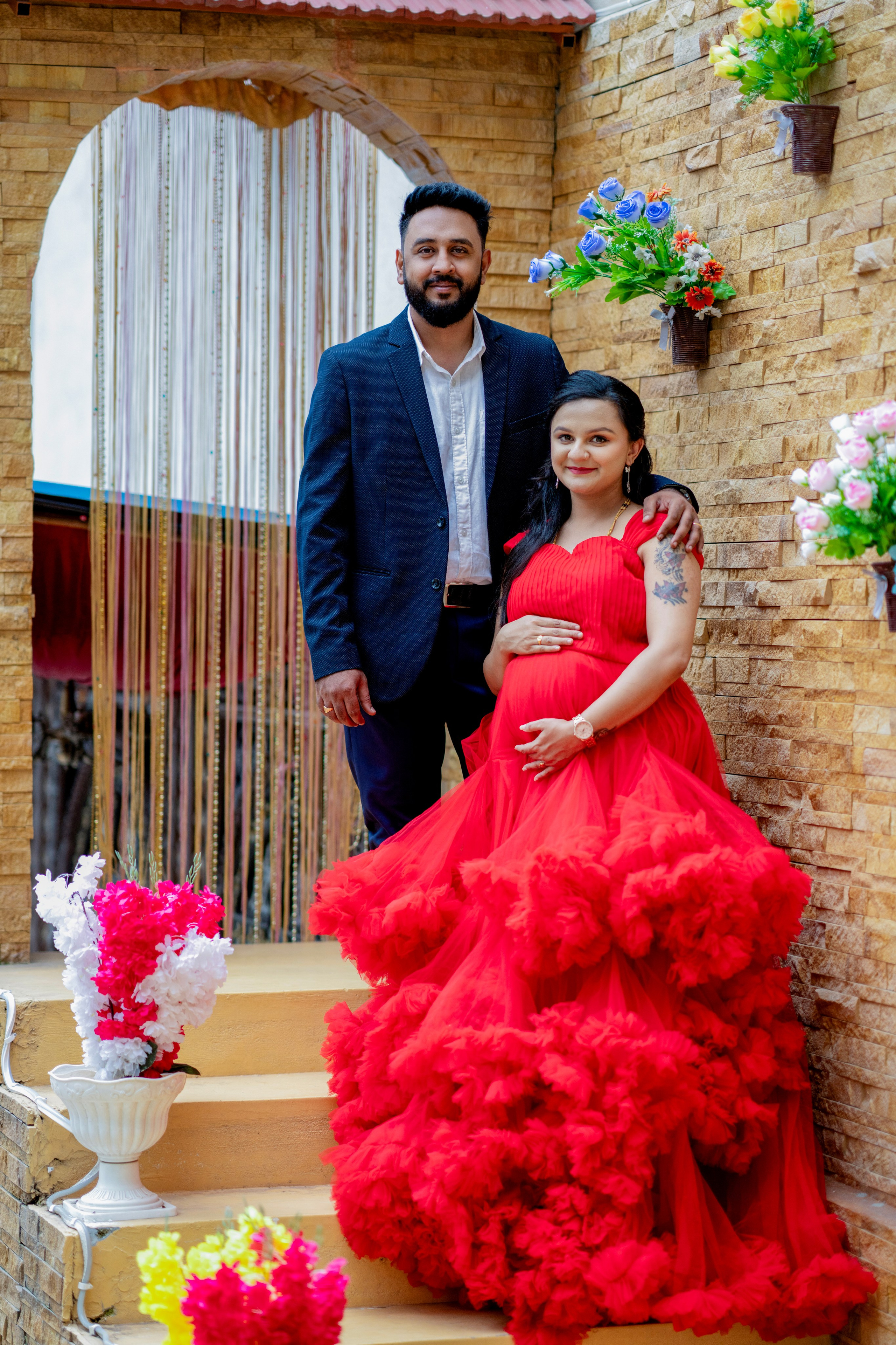 Maternity photoshoot in Bengaluru featuring a woman in a voluminous red ruffled gown and a man in a navy suit posing on stairs against a stone wall with flowers.