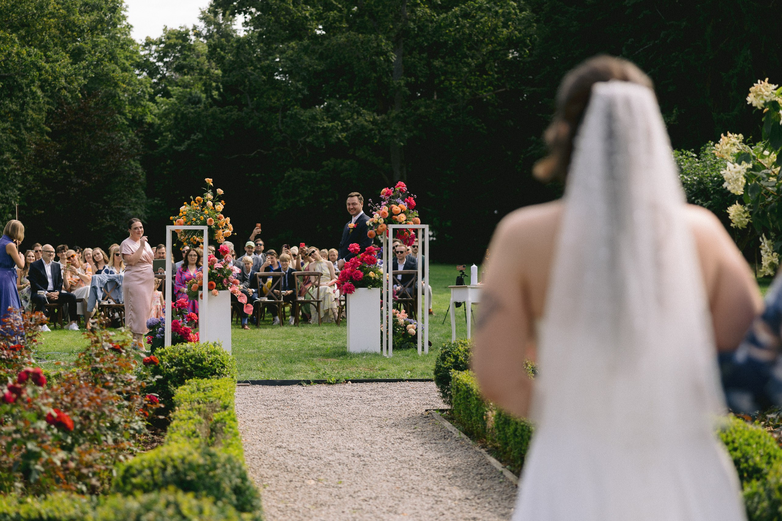 Līgava ar plīvuru dodas pa dārza celiņu pretī ceremonijas vietai.