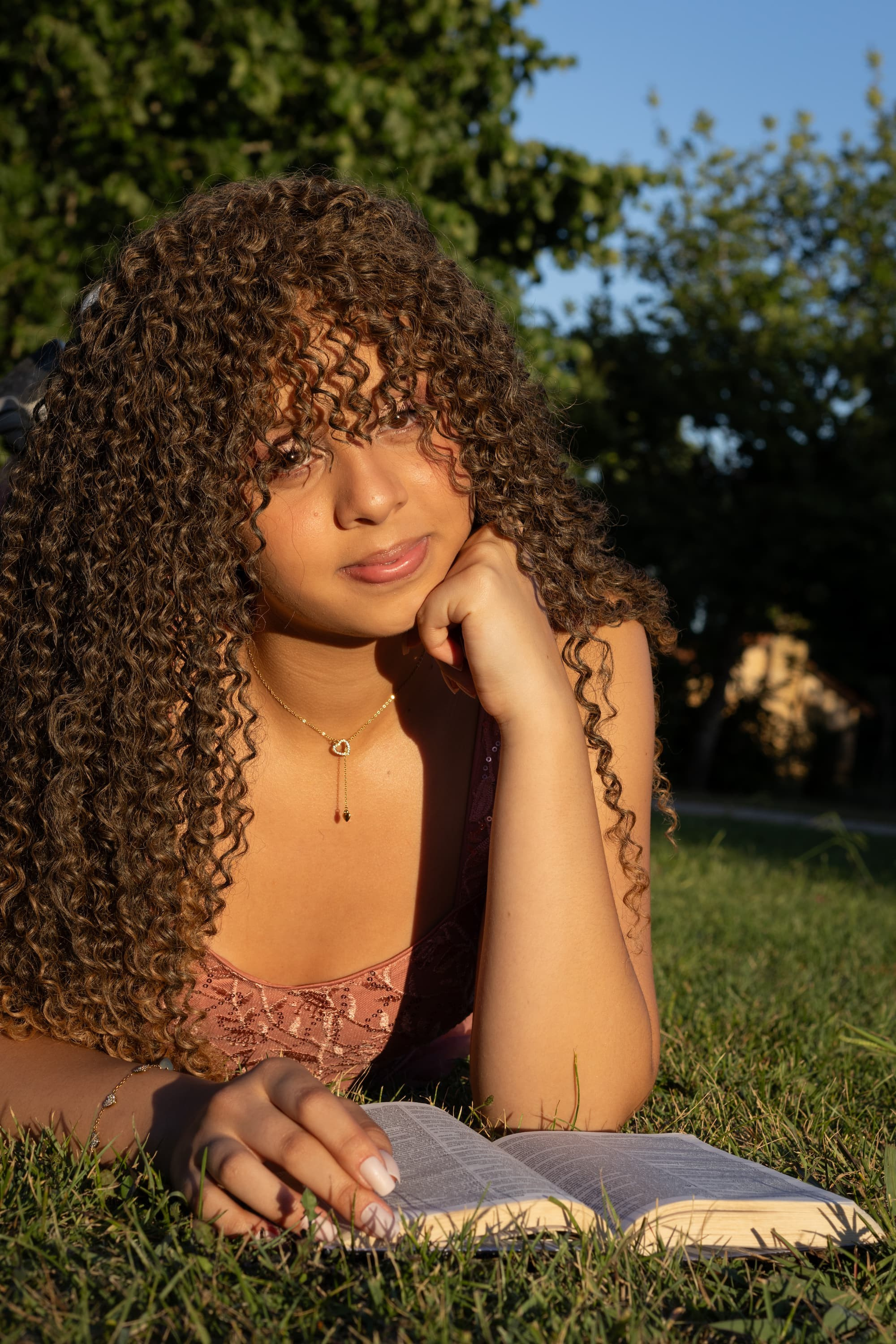 Close-up de mulher com cabelos cacheados dourados iluminados pelo sol, inclinada sobre um livro aberto na grama. Foco na iluminação dourada (golden hour).